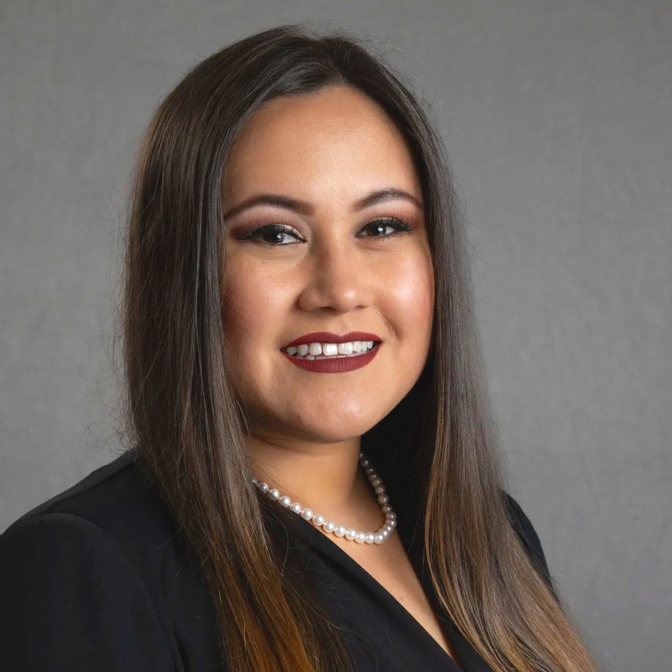 Professional woman with long wavy brown hair, wearing a maroon blazer and cream-colored blouse, smiling at camera against a dark gray background.
