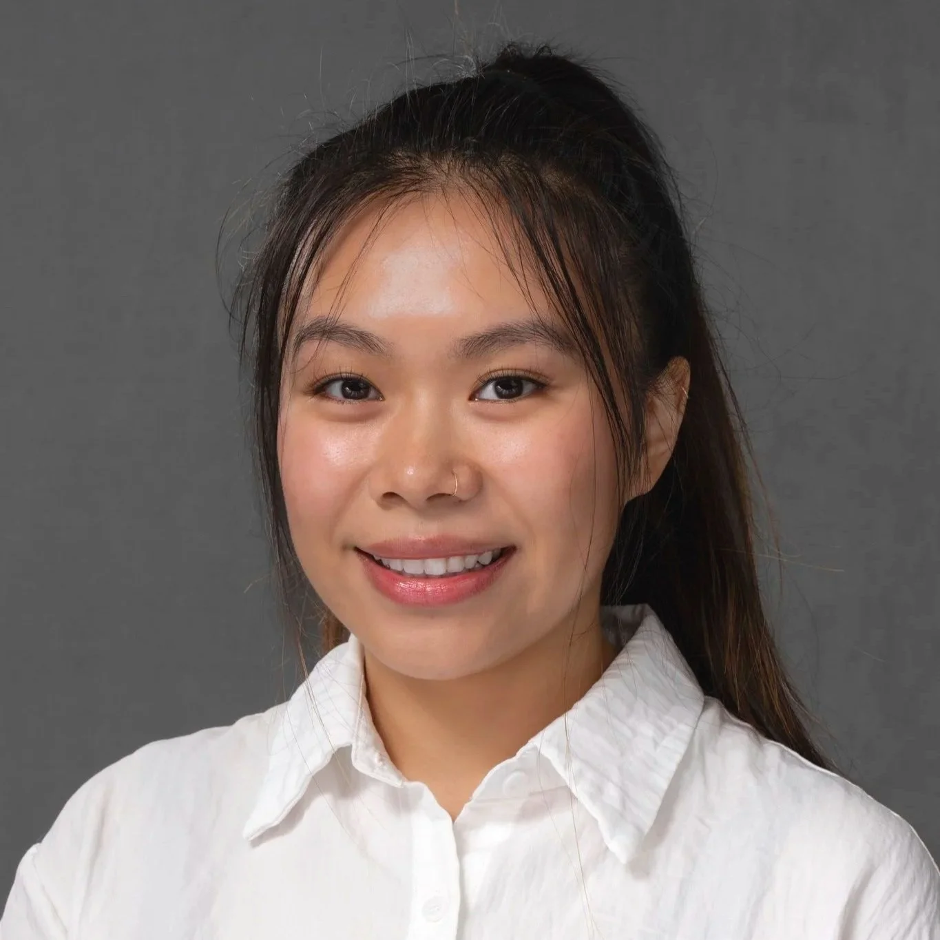 Portrait of a smiling young Asian woman with long brown hair, wearing a white top, a red string necklace with a green pendant, and two layered necklaces including one with a lightning bolt charm, with a tattoo of a flamingo and flowers on her shoulder against a dark background.
