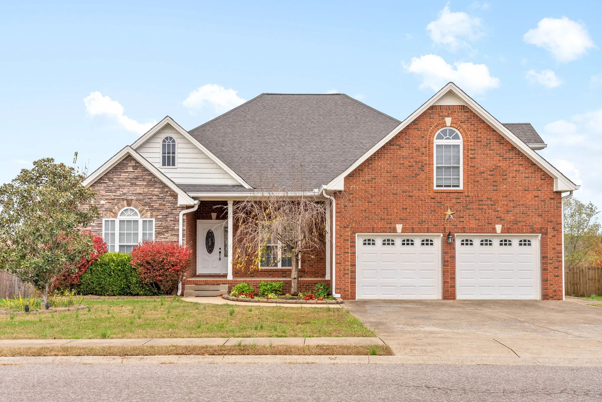 Front view of a two-story house with brick and stone exterior, white garage doors, a concrete driveway, a small front porch with steps, and landscaping including trees and bushes under a partly cloudy sky.