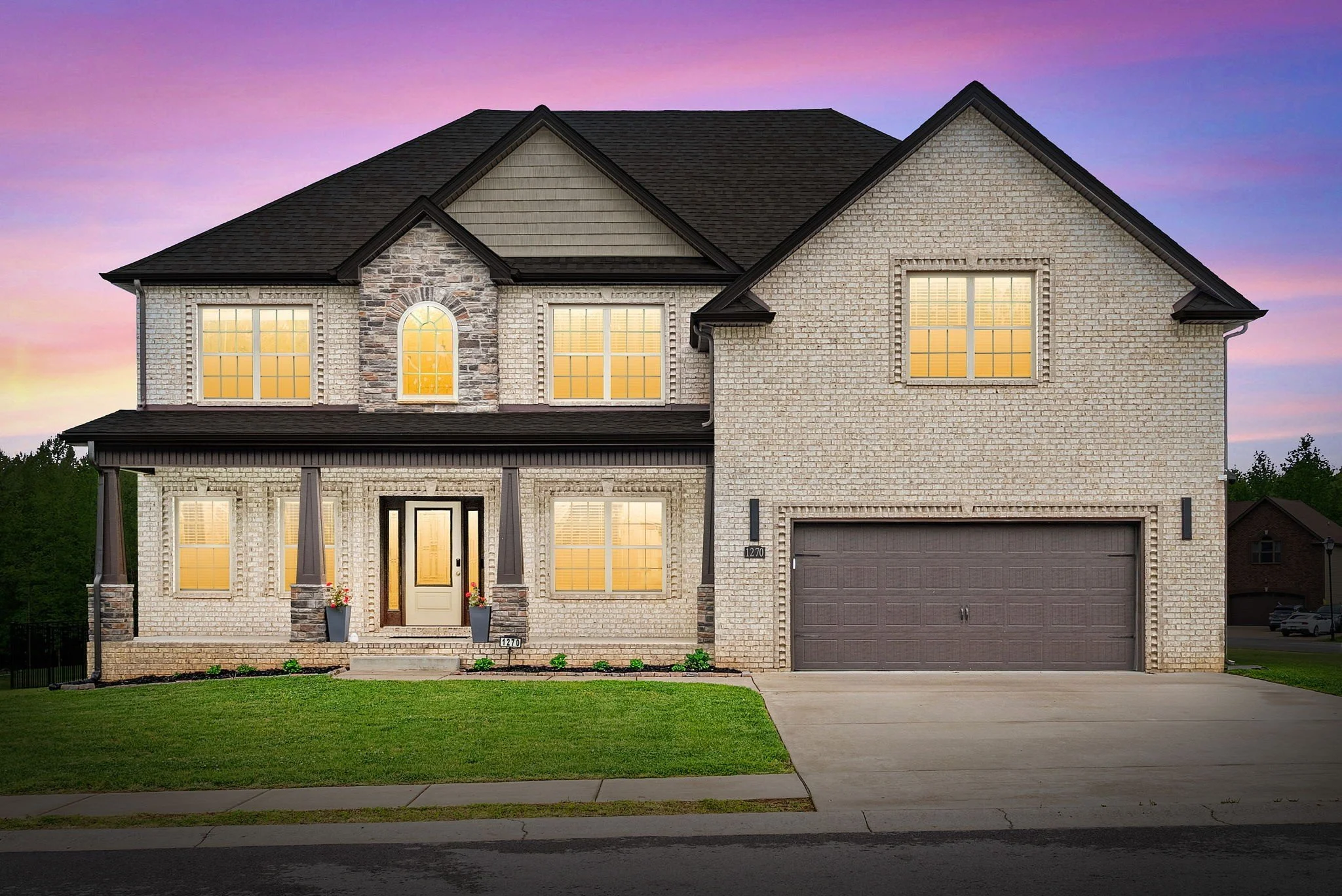 A two-story brick house with a black front door, black shutters, and large windows with white frames, situated under a sunset sky with orange and pink clouds.