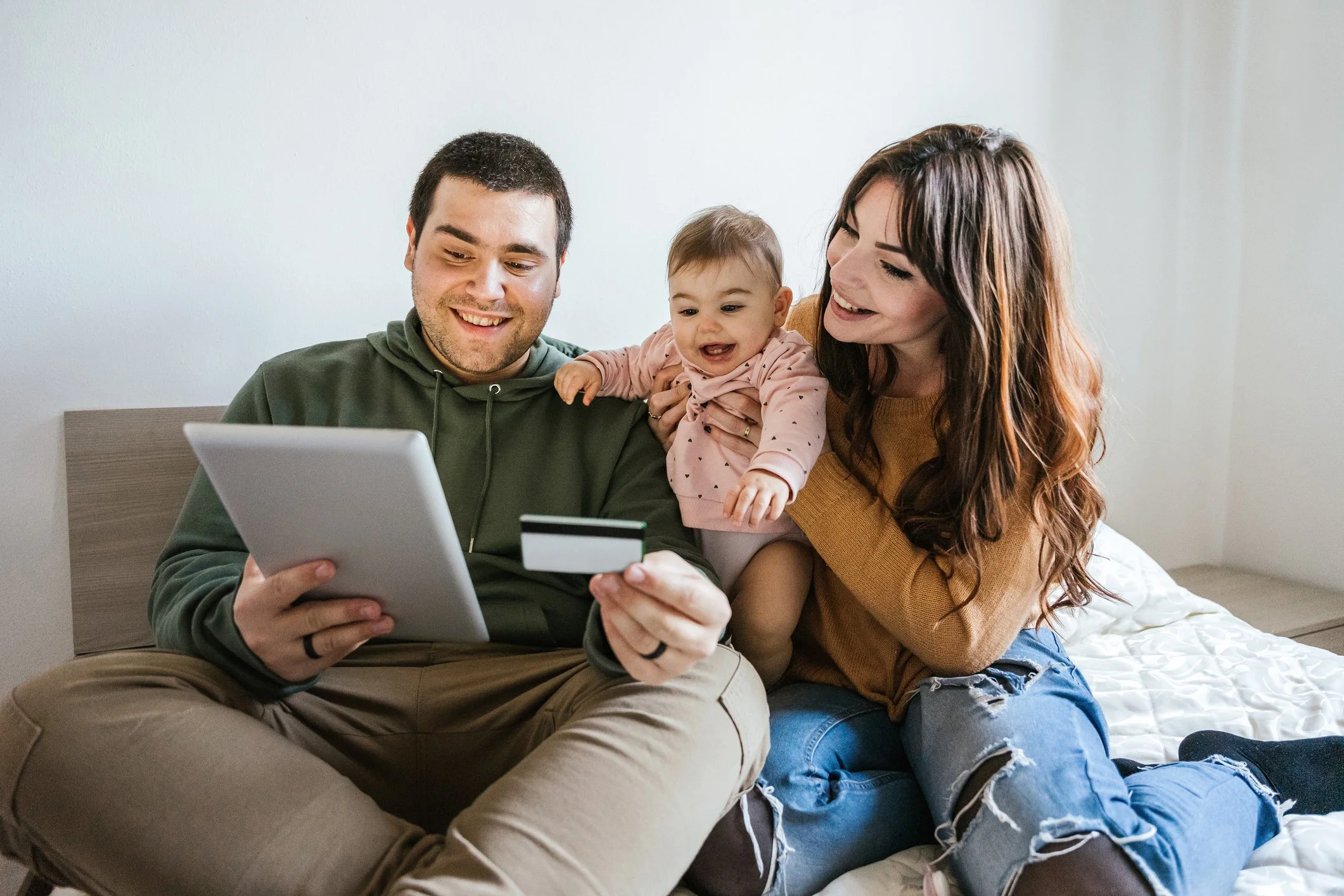 Family of three sitting on a bed looking at a tablet and a credit card, smiling and enjoying their time together.