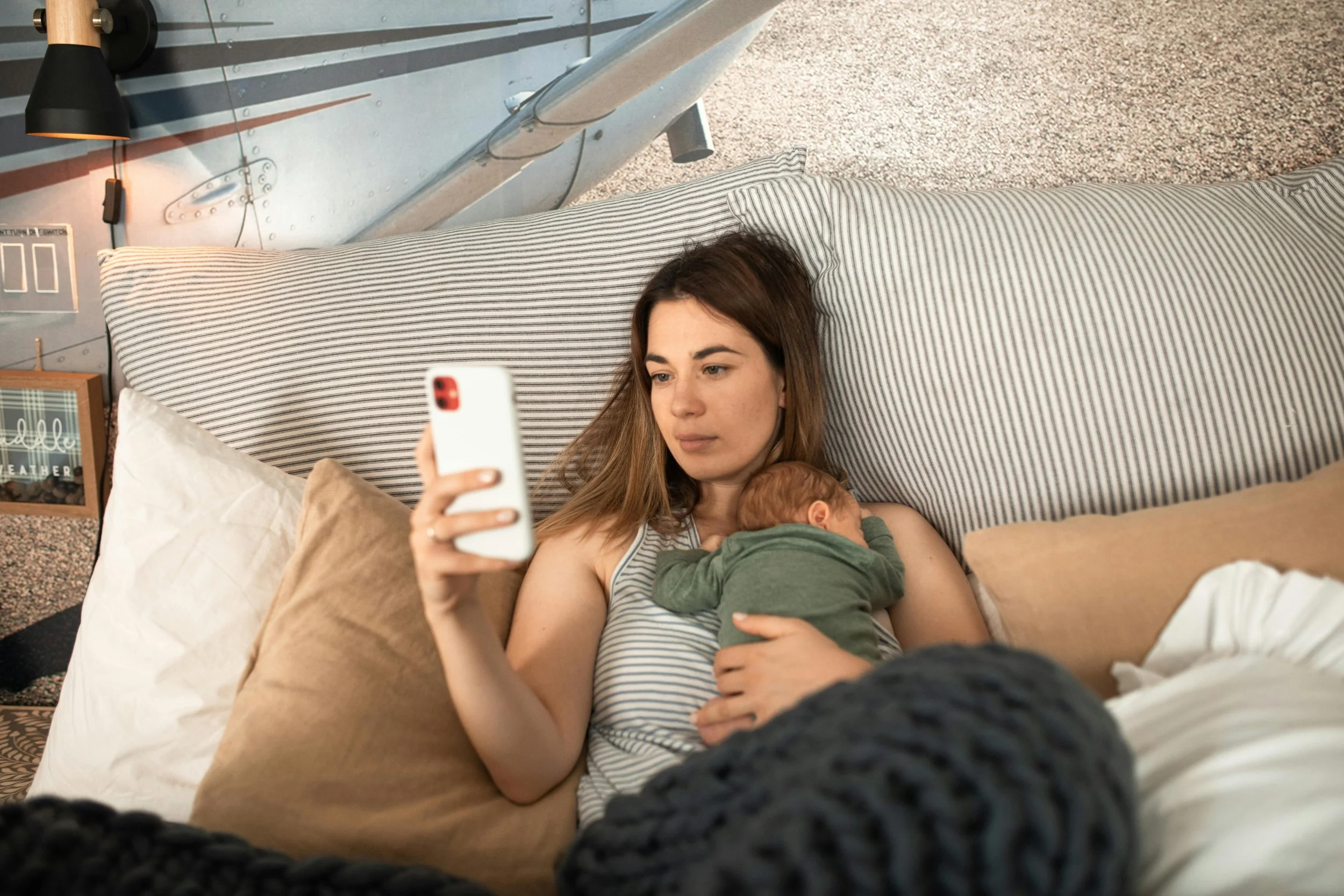 A woman is lying on a bed with a baby nursing, holding a smartphone, in a bedroom with striped pillows and a nautical-themed wall.