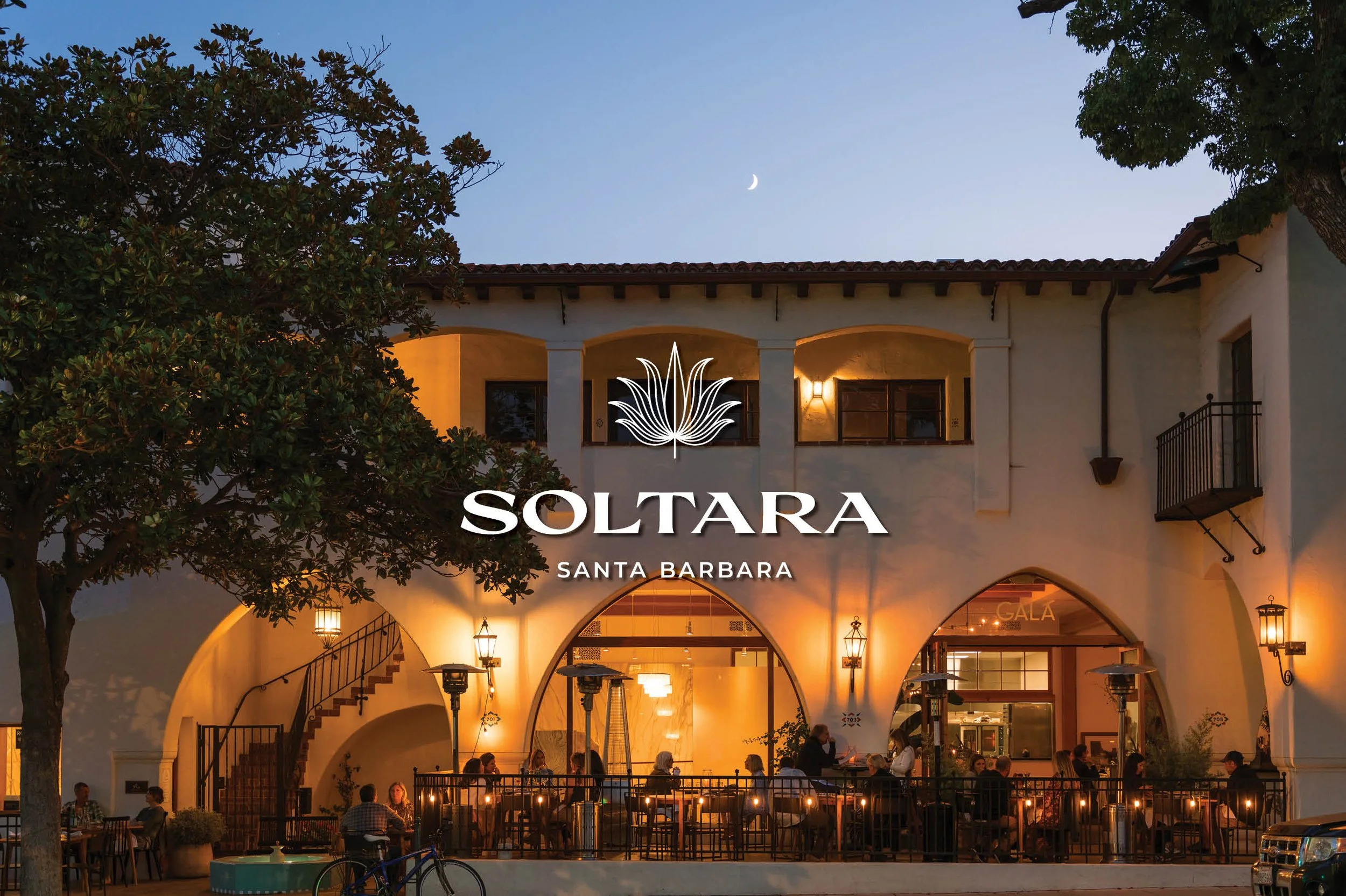 Exterior view of Soltara restaurant in Santa Barbara during evening, with outdoor dining, warm lighting, trees, and a crescent moon in the sky.