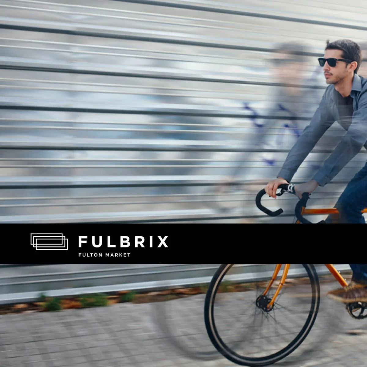 A man wearing sunglasses and a gray shirt riding a yellow bicycle with black wheels past a metallic fence, with a black banner across the middle that reads "FULBRIX FULTON MARKET".