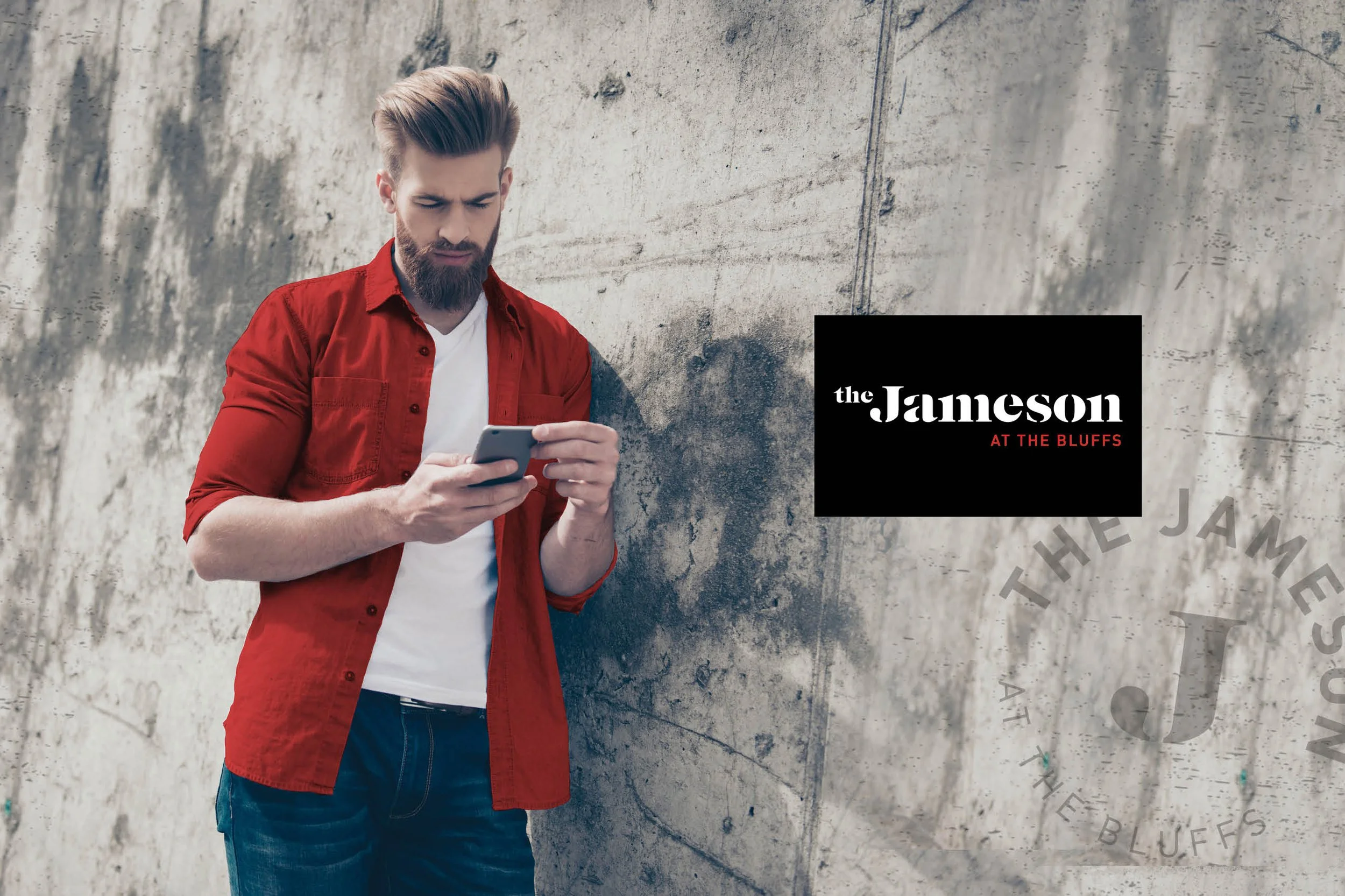 Young man with a beard wearing a red shirt over a white t-shirt, looking at his phone while leaning against a concrete wall. There is a black rectangular sign to his right with white and red text that reads 'the Jameson at the Bluffs.'