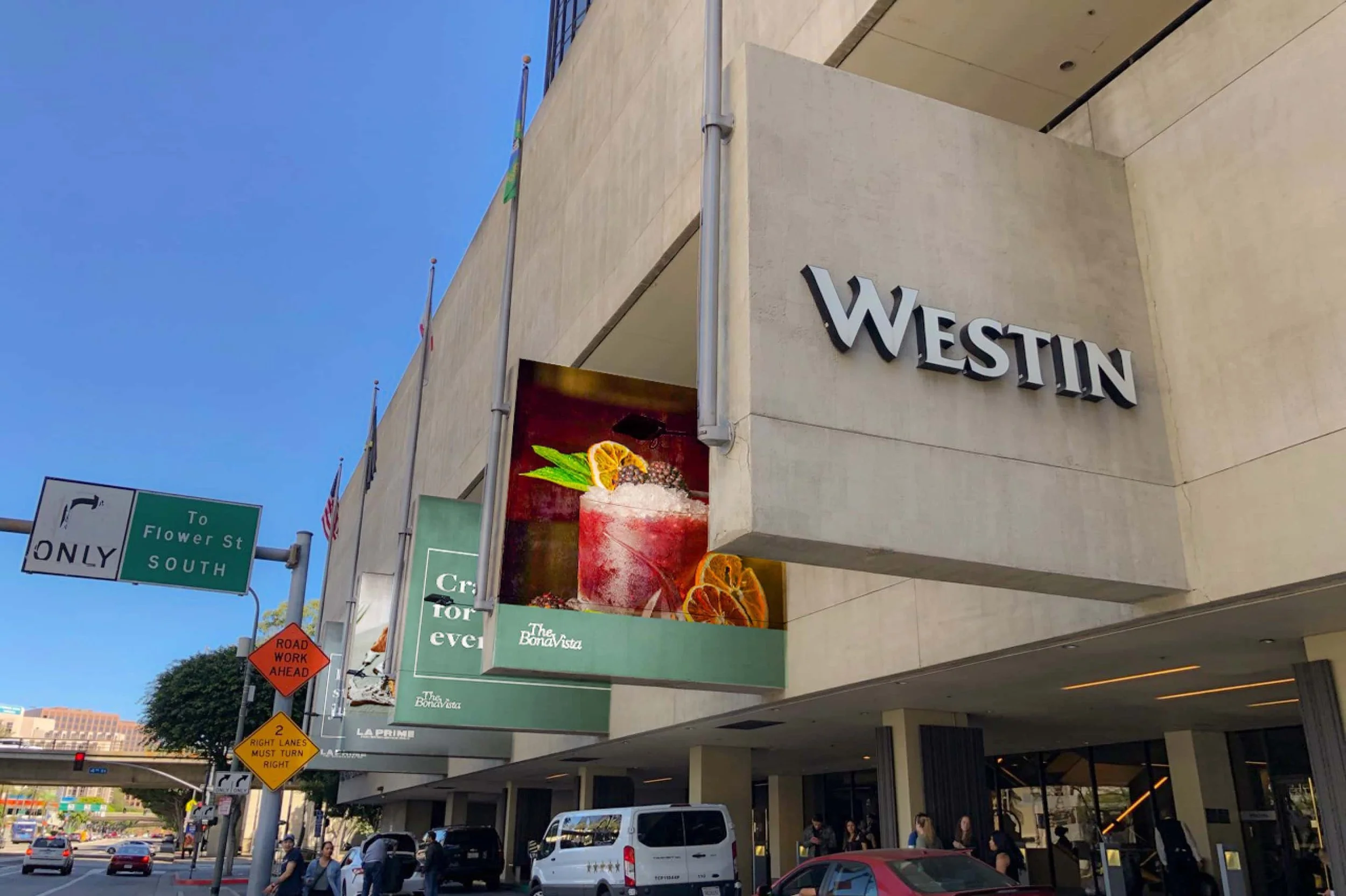 Front view of Westin hotel with a large sign, adjacent digital billboard displaying a cocktail with fruit garnishes, street signs, traffic, and people at the sidewalk.