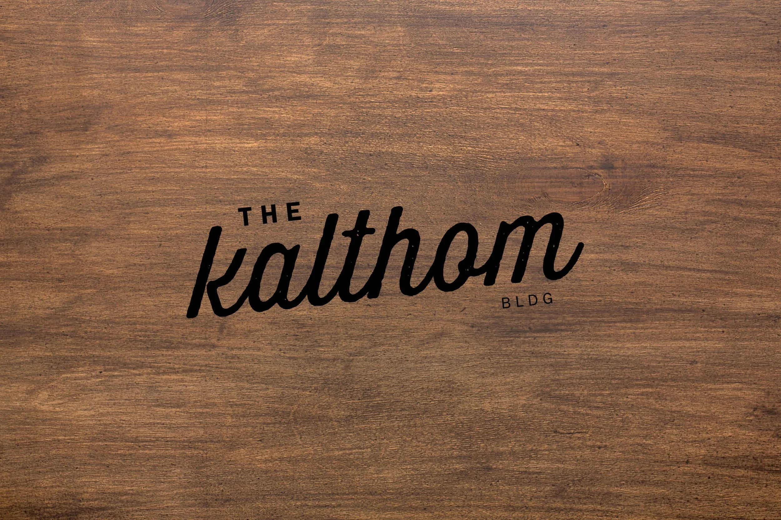 Wooden sign with black text reading 'The Kalthom BLDG' in a decorative font. Logo design by Richard Shanks.
