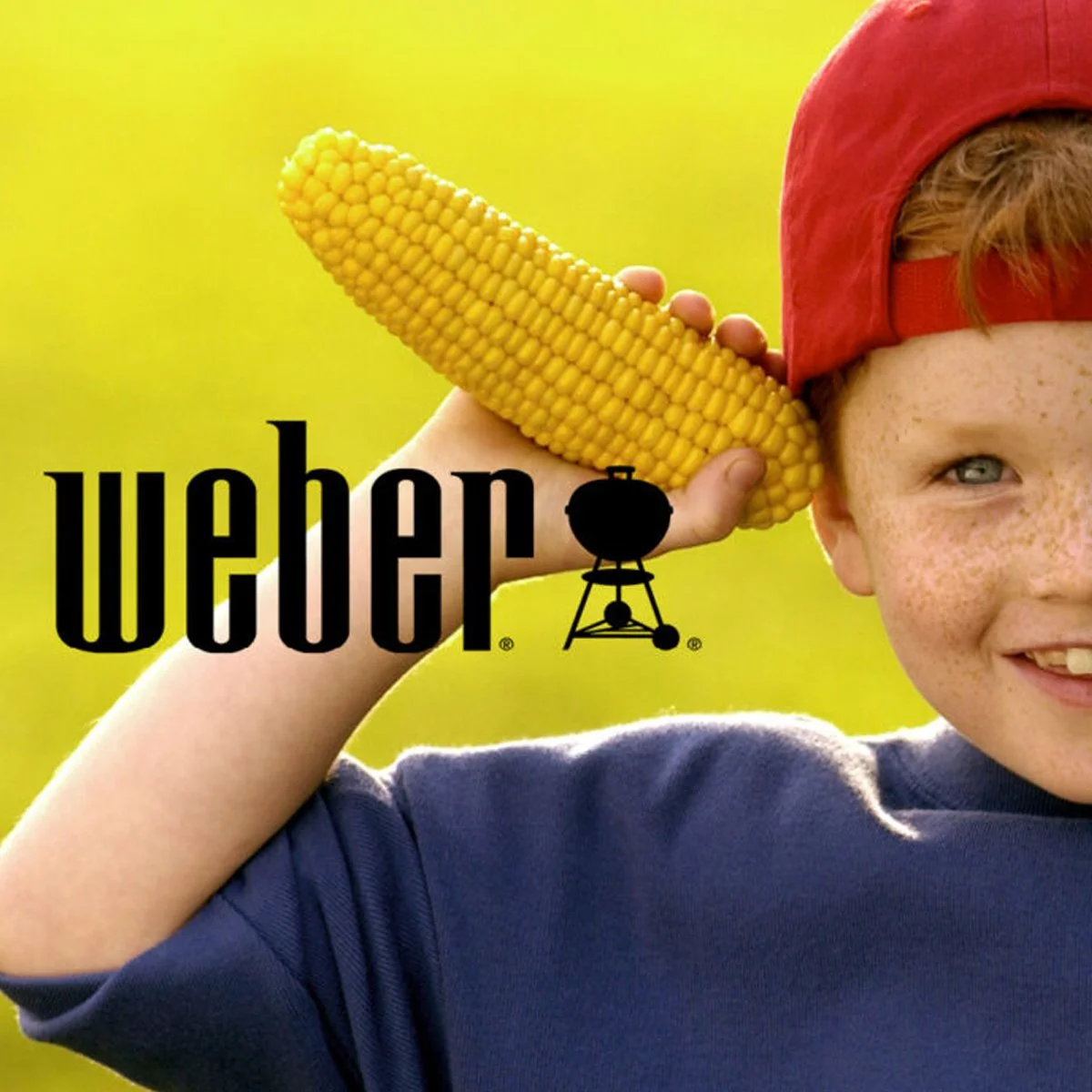 Young boy holding a cob of corn to his ear like a headphone, wearing a red cap backwards, smiling, and standing outdoors