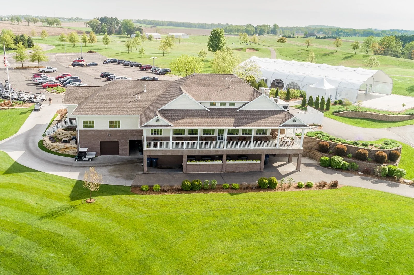 Aerial view of a golf clubhouse with a parking lot, surrounded by greenery and a large white tent structure nearby.
