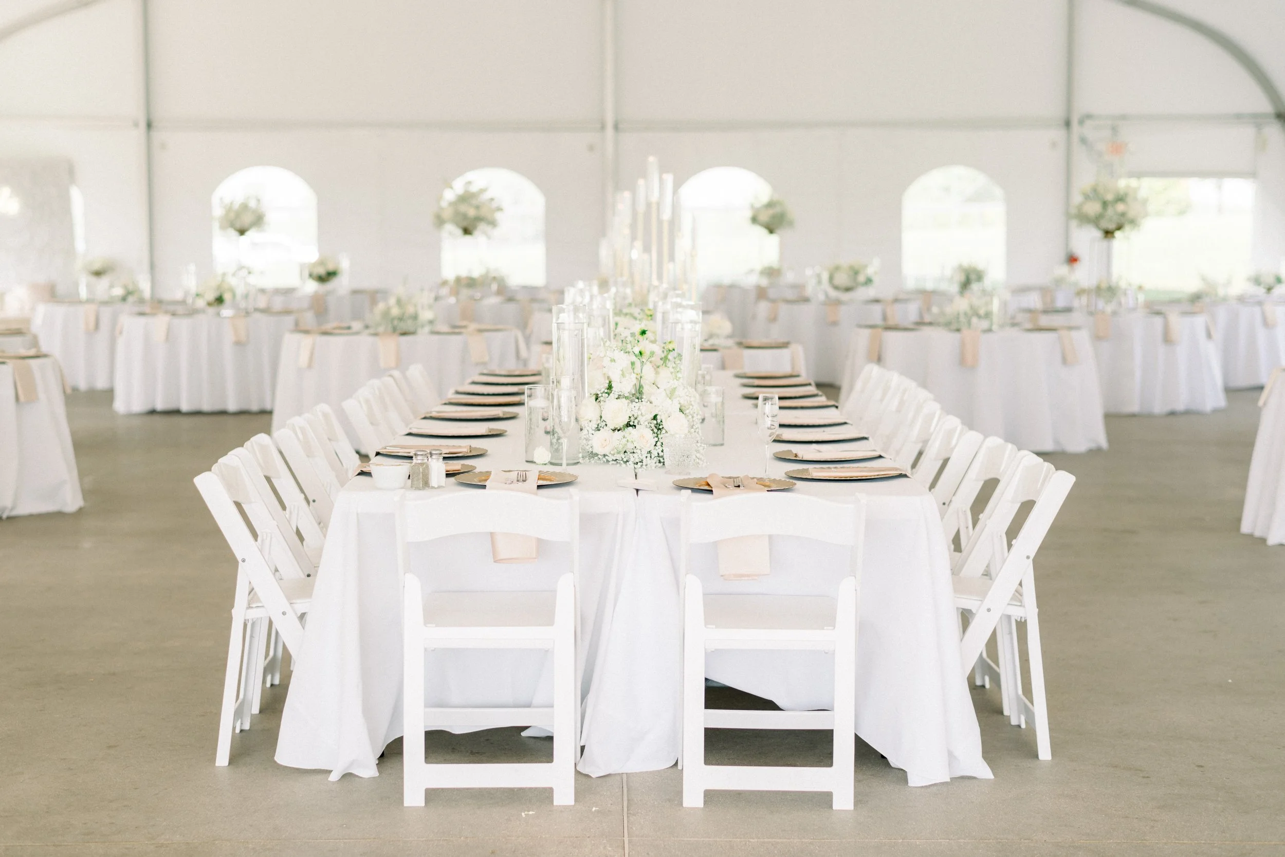Elegant banquet table setting with white tablecloths, white chairs, and floral centerpieces in a large reception hall.