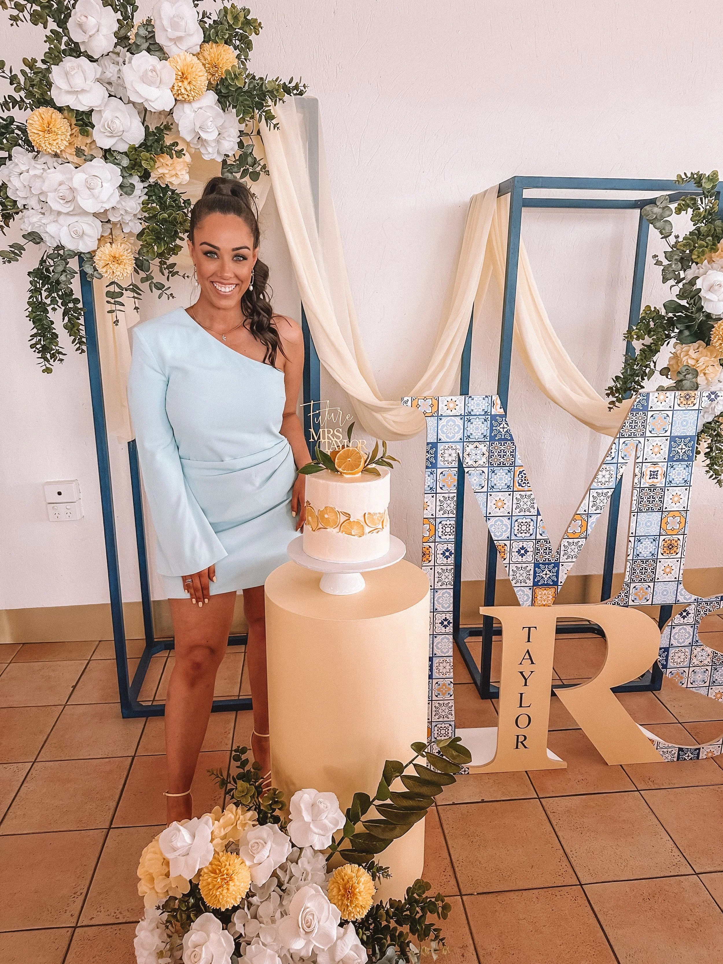 A woman in a light blue dress standing next to a small cake with lemon decorations, decorated with a floral arrangement in front and a wedding celebration backdrop with large letters spelling 'M' and 'R' and the name 'Taylor'.
