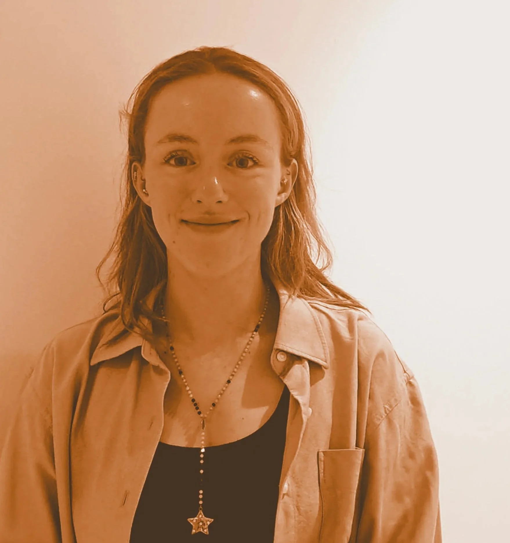 Jeune femme aux cheveux roux et yeux marron, portant un t-shirt noir et une veste beige, avec un collier à breloque en forme d'étoile, se tenant devant un mur blanc.