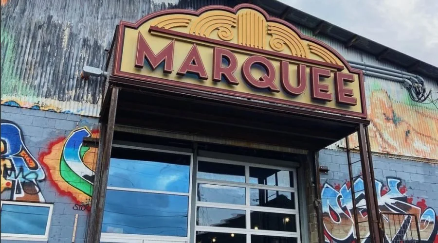 The exterior of a building with a large sign reading "MARQUEE" above the entrance, and colorful graffiti art on the brick wall to the side.