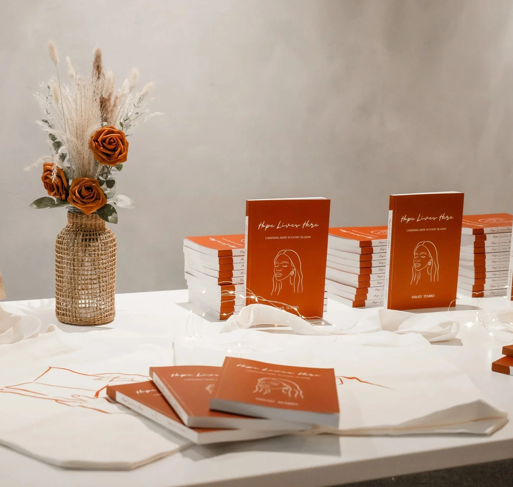 Table with books, a price list, a decorative vase with roses, and tote bags.