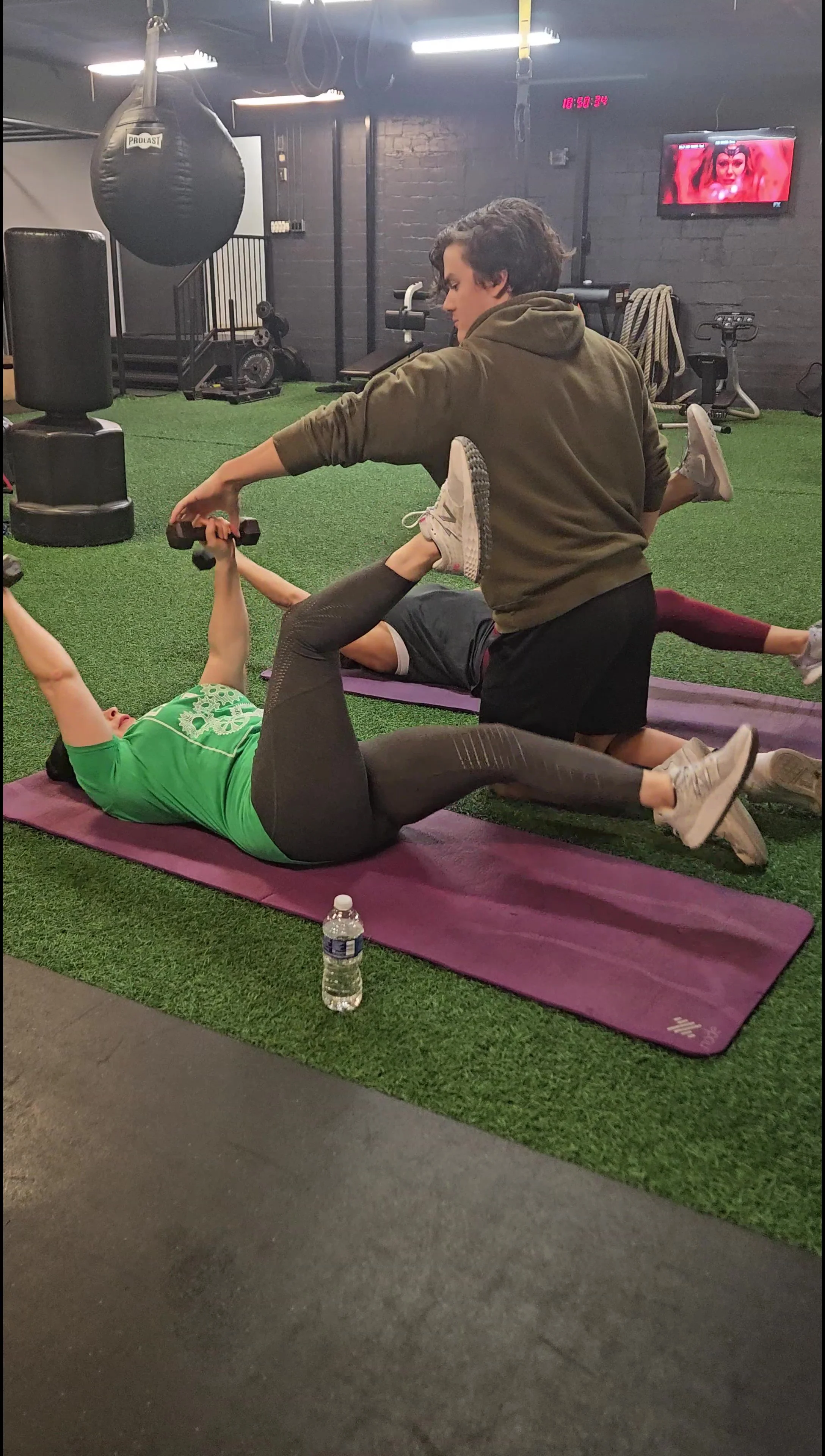 A woman in a green shirt and black leggings lying on a purple exercise mat doing a workout with dumbbells, assisted by a trainer in a brown hoodie. There is a water bottle in front of the woman on the floor. Other people are exercising on mats in the background, and gym equipment such as a punching bag, a large speed bag, and a television on the wall are visible.