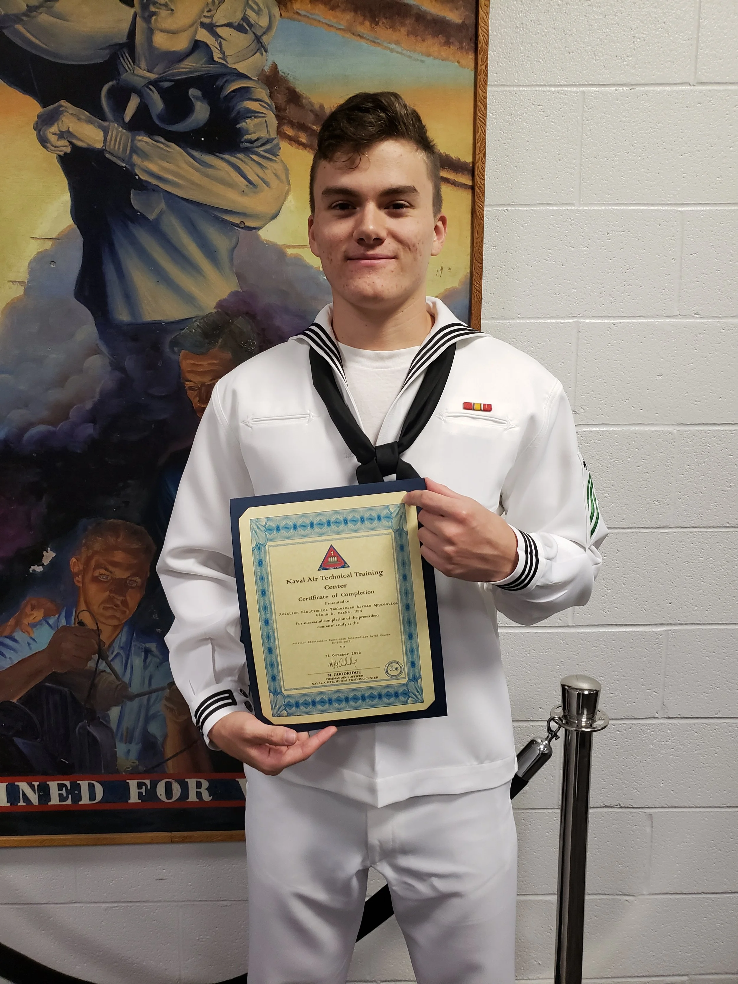 A young man in a white military uniform holding a naval air technical training certificate, standing in front of a colorful mural depicting sailors and ships.