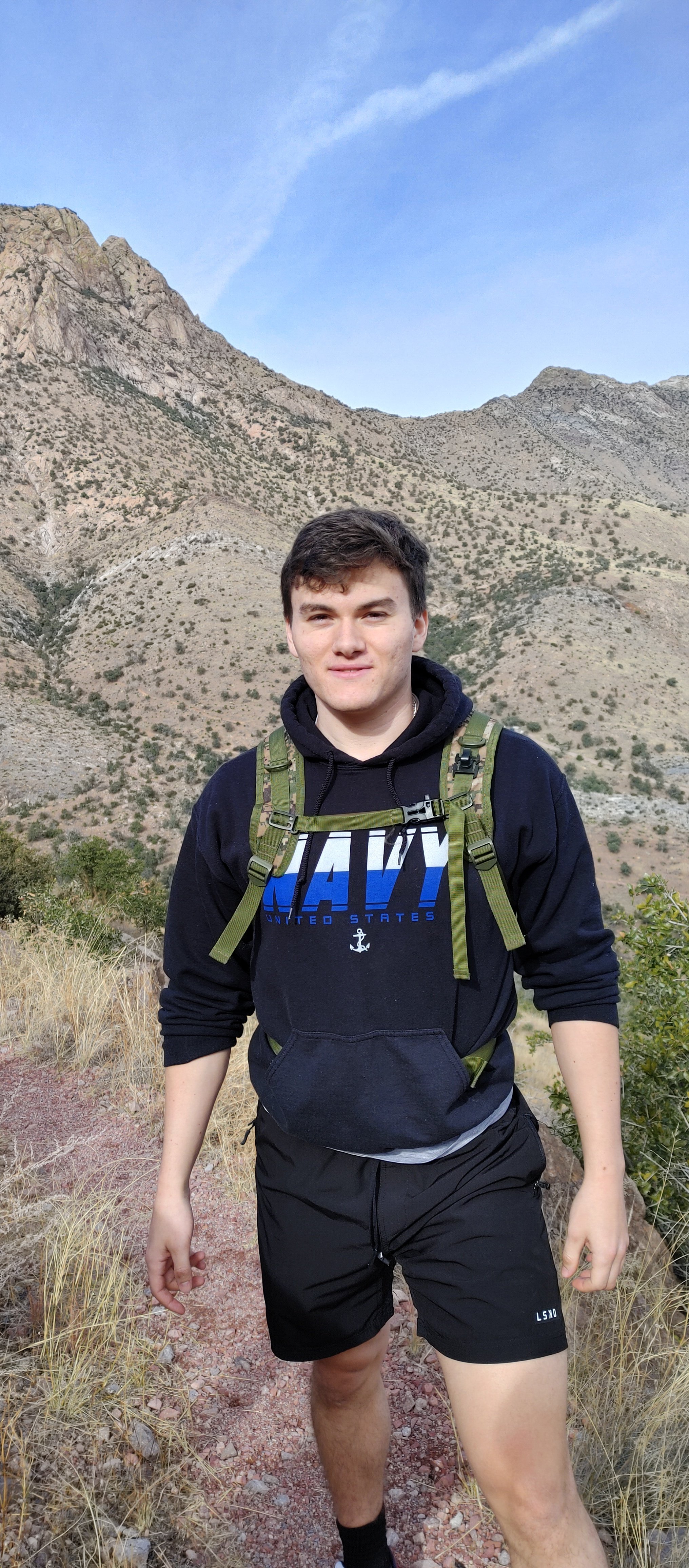 Young man in black hoodie and shorts hiking in mountainous terrain with a backpack, dry grass, and clear sky in the background.