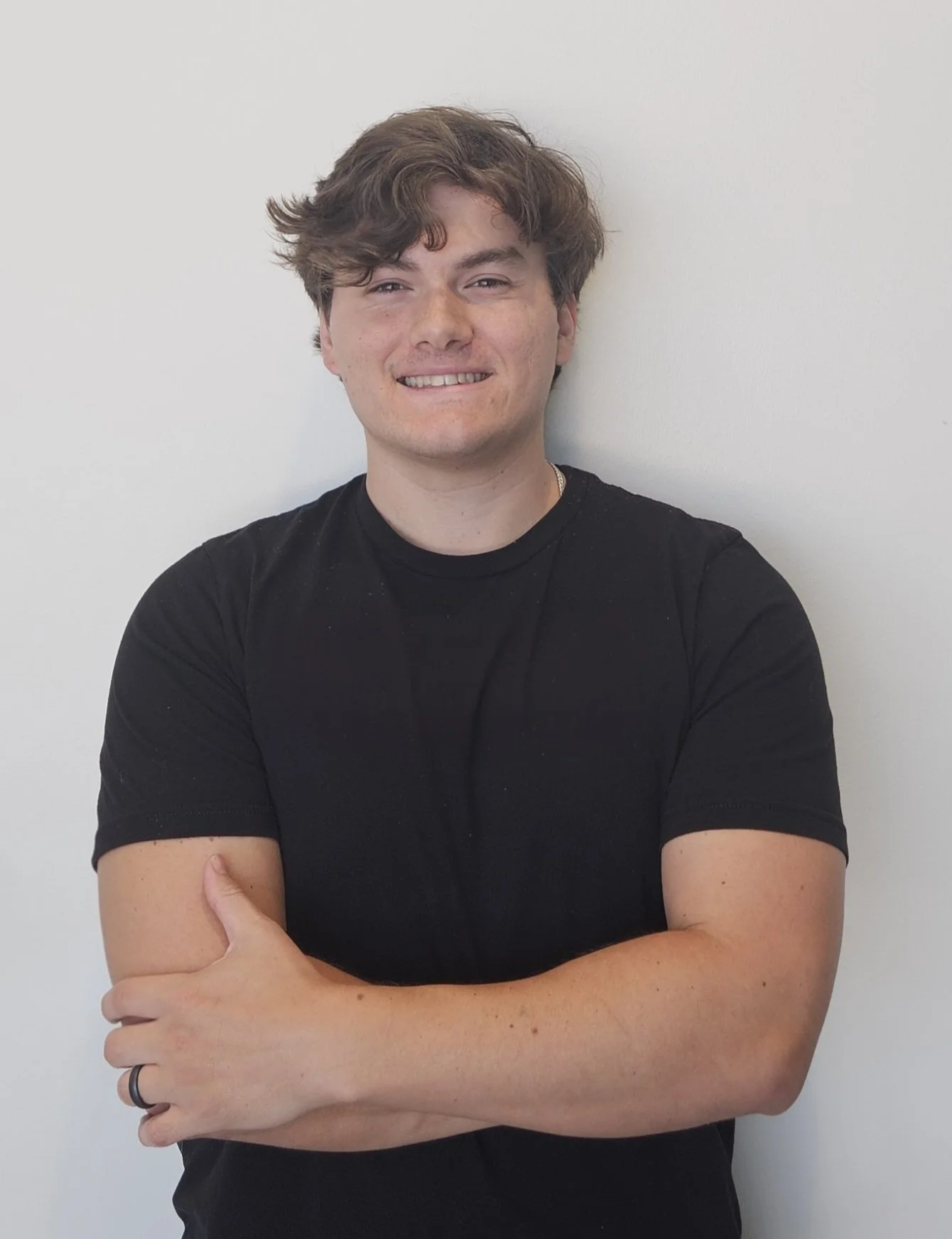 A young man with wavy brown hair, wearing a black t-shirt, smiling, with arms crossed, standing against a plain white wall.