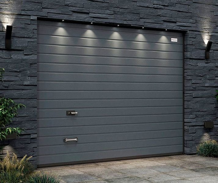 Un garage avec porte en métal gris dans un mur en pierre noir, éclairé par des lampes encastrées.