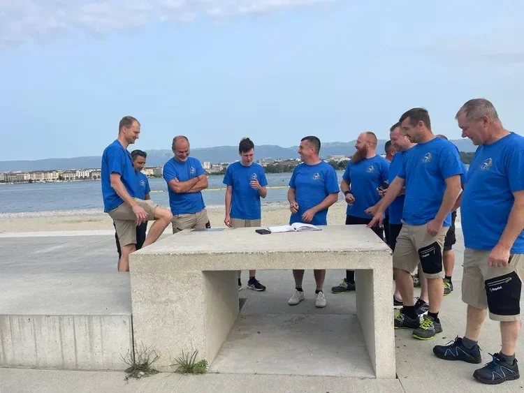 Groupe de personnes en t-shirts bleus autour d'une table en extérieur près d'un lac, probablement lors d'une réunion ou d'un événement.