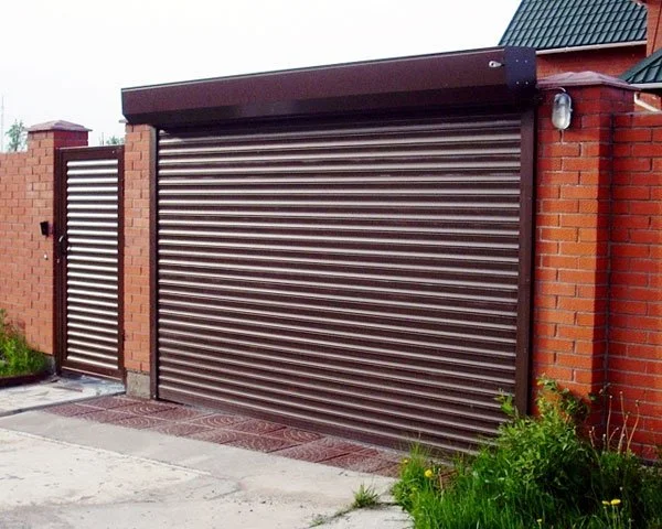 Grand portail de garage en métal avec une petite porte adjacente, entouré de murs en briques rouges, avec des lampes extérieures et un toit en tuiles vertes.