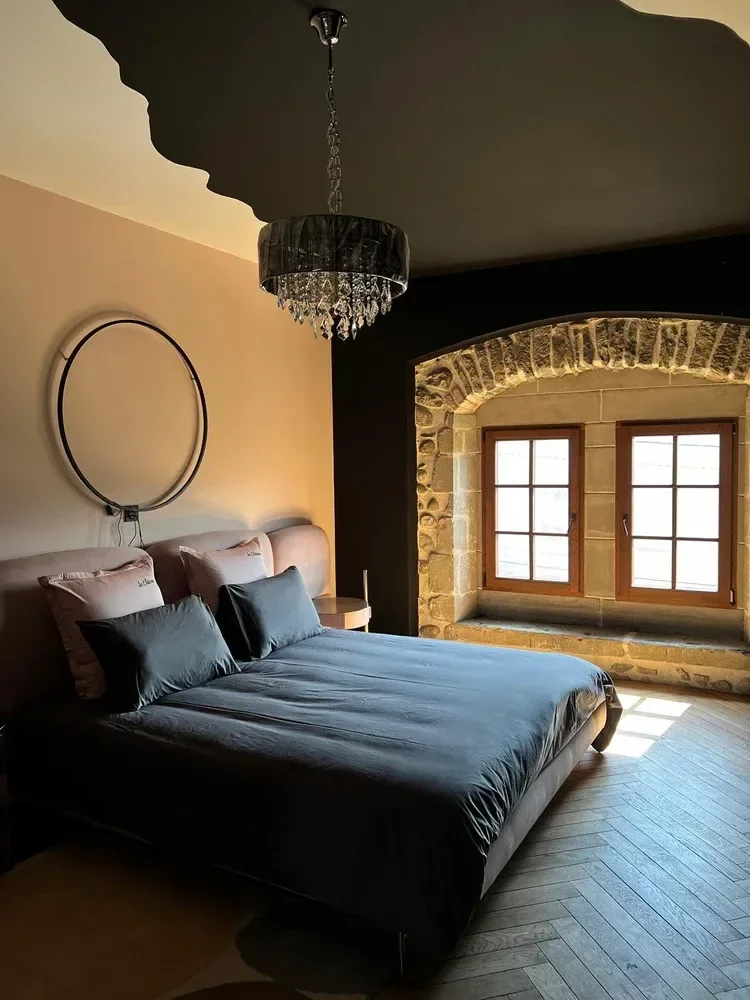 Chambre à coucher avec un lit au lit avec des draps gris foncé, deux oreillers gris et deux oreillers rose clair, un plafond noir avec un lustre en cristal et un mur beige à gauche. Il y a une fenêtre avec une pierre ancienne et un cadre en bois.