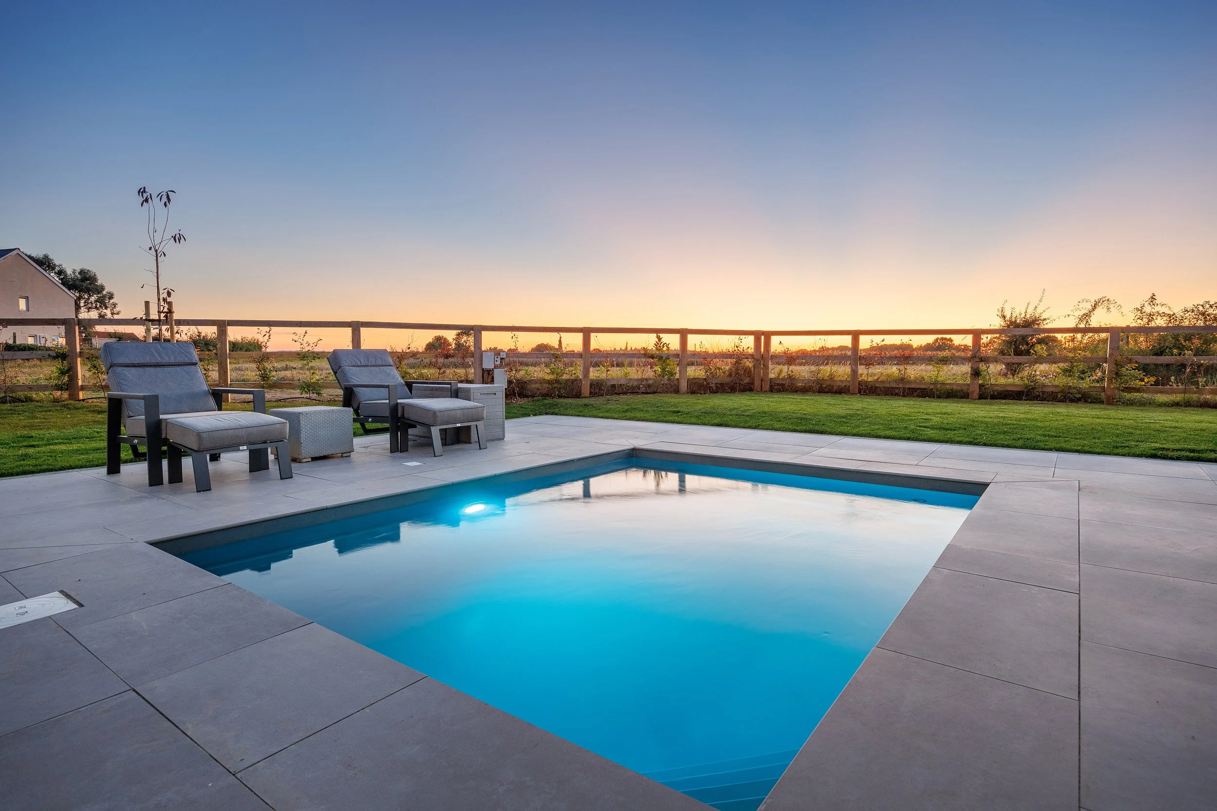 Residential backyard with a swimming pool, lounge chairs, and a wooden fence at sunset.
