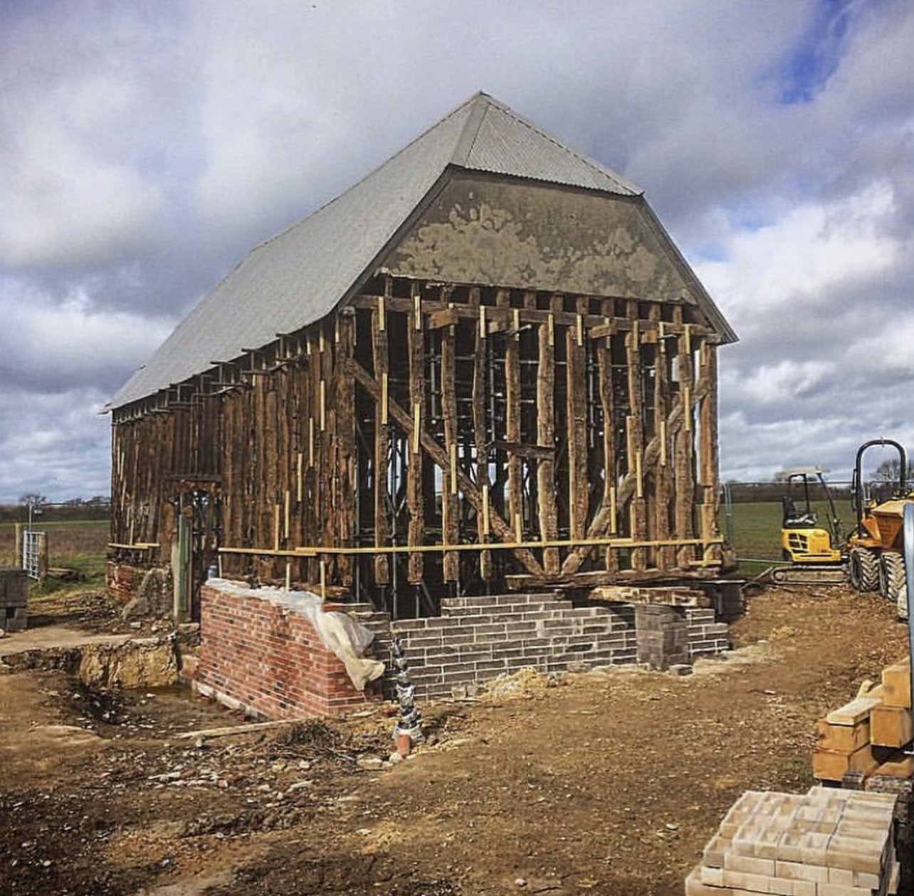 Wooden barn under construction with visible framing, brick foundation partially built, and construction equipment nearby on a dirt lot surrounded by open fields.
