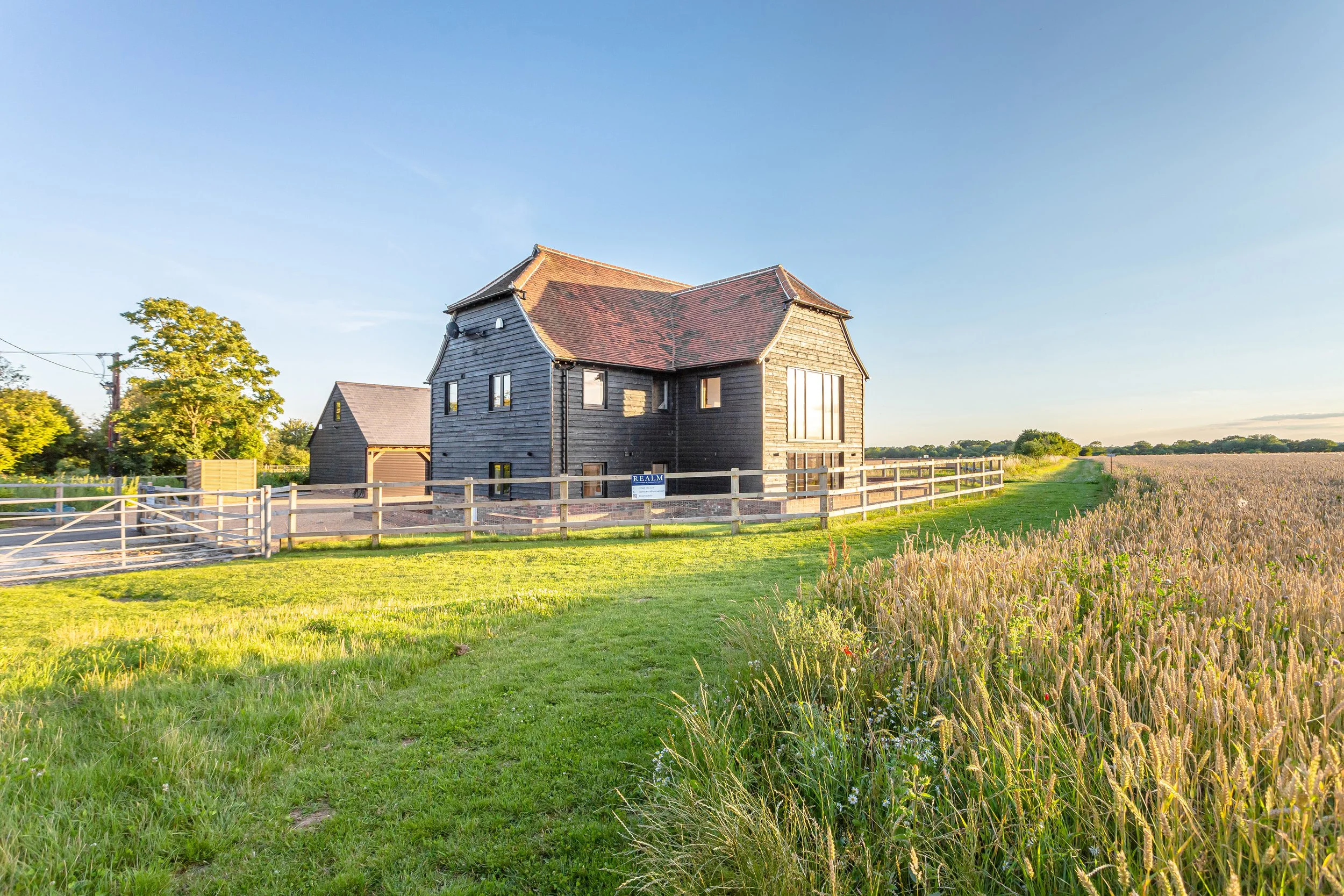 A large black wooden house with a red tile roof on a farm, surrounded by green grass and a wheat field, under a clear blue sky.