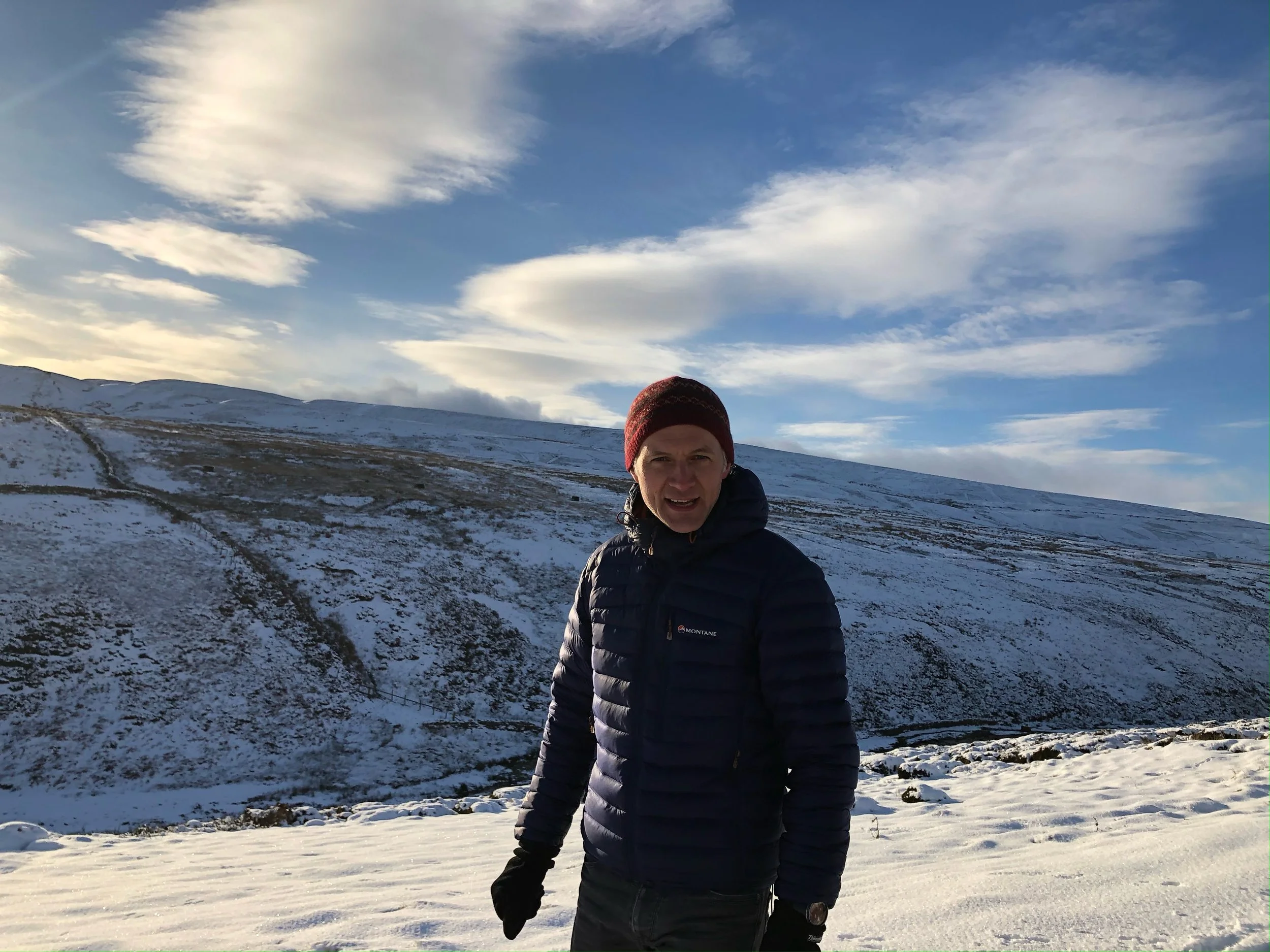 A man wearing a dark blue jacket and a red beanie stands outdoors in a snowy landscape with rolling hills. The sky is partly cloudy with patches of blue visible.