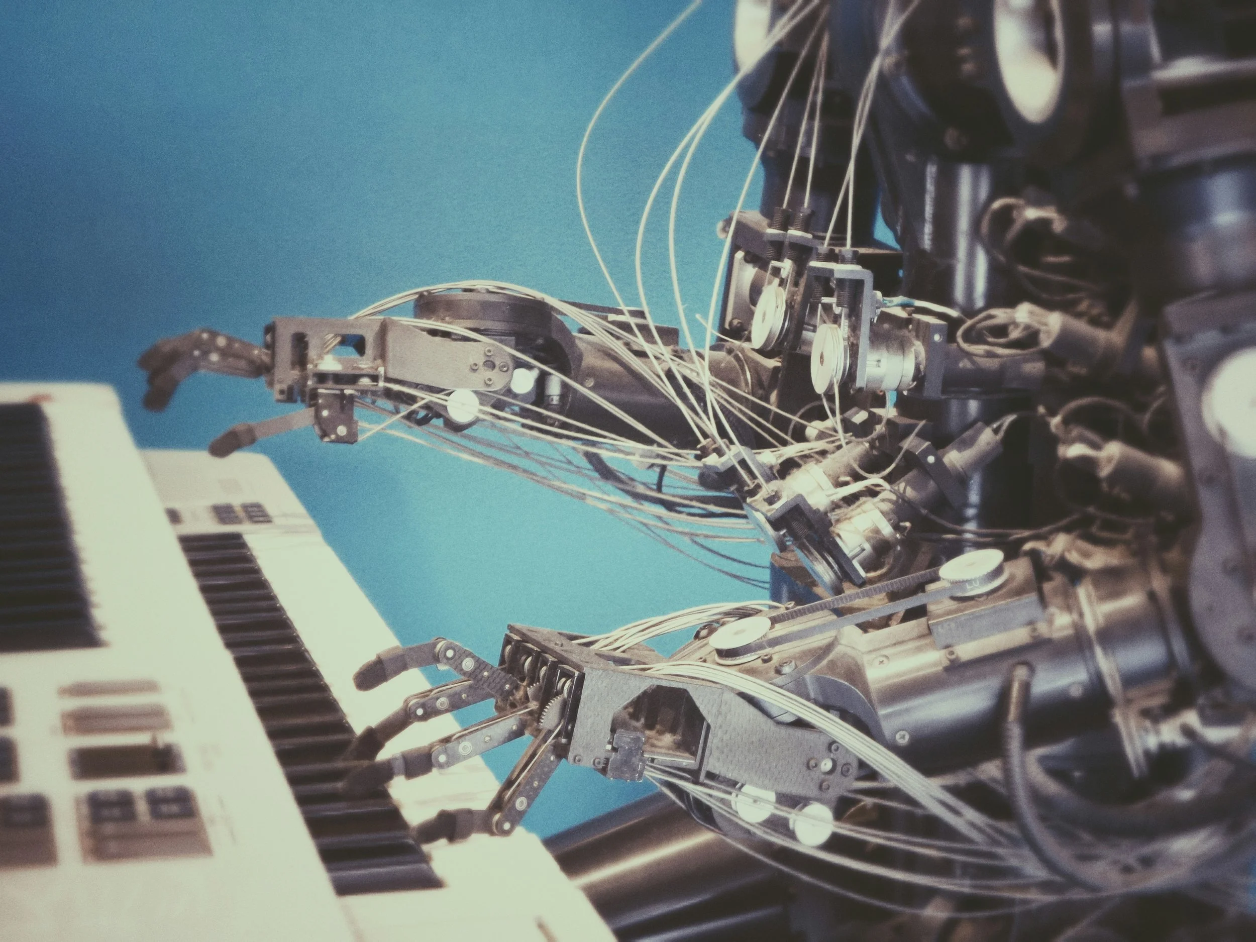 Close-up of a robot with mechanical arms and wires playing a keyboard against a blue background.