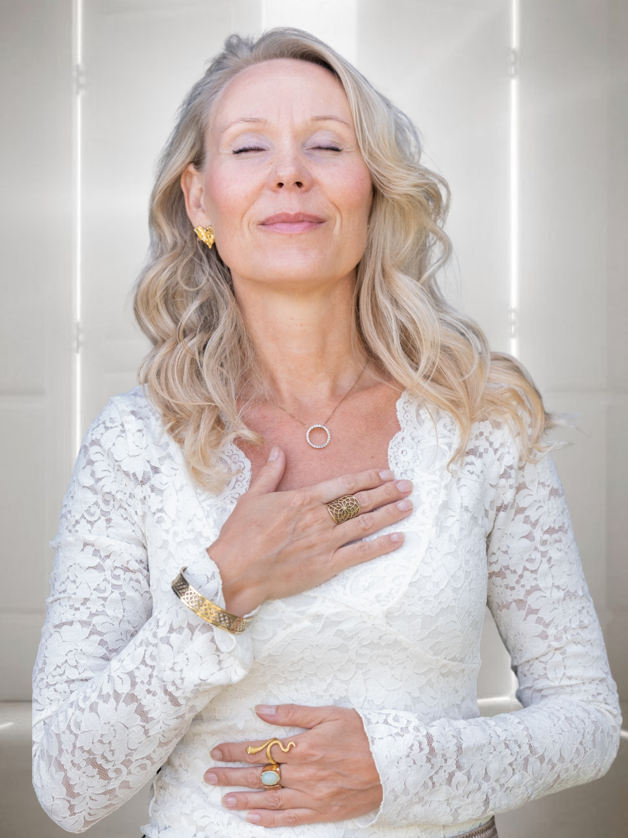 En glad kvinde med lukkede øjne, iført hvid blondekjole med fine smykker, der står foran en lys baggrund.
