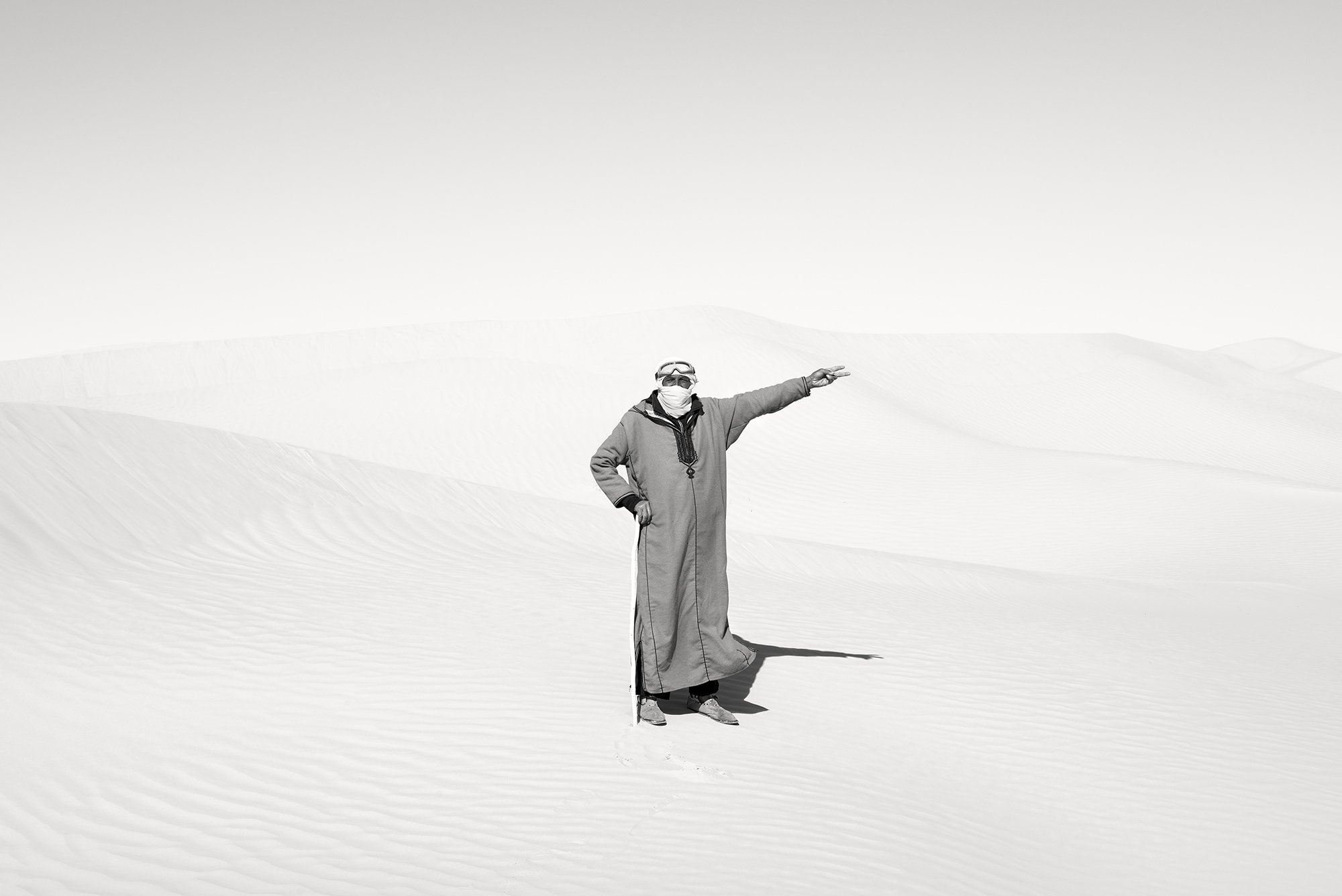High-key black and white portrait of a man in traditional desert dress and goggles, standing in a bright, white sand dune landscape with one arm outstretched, by Isabelle Kettner.
