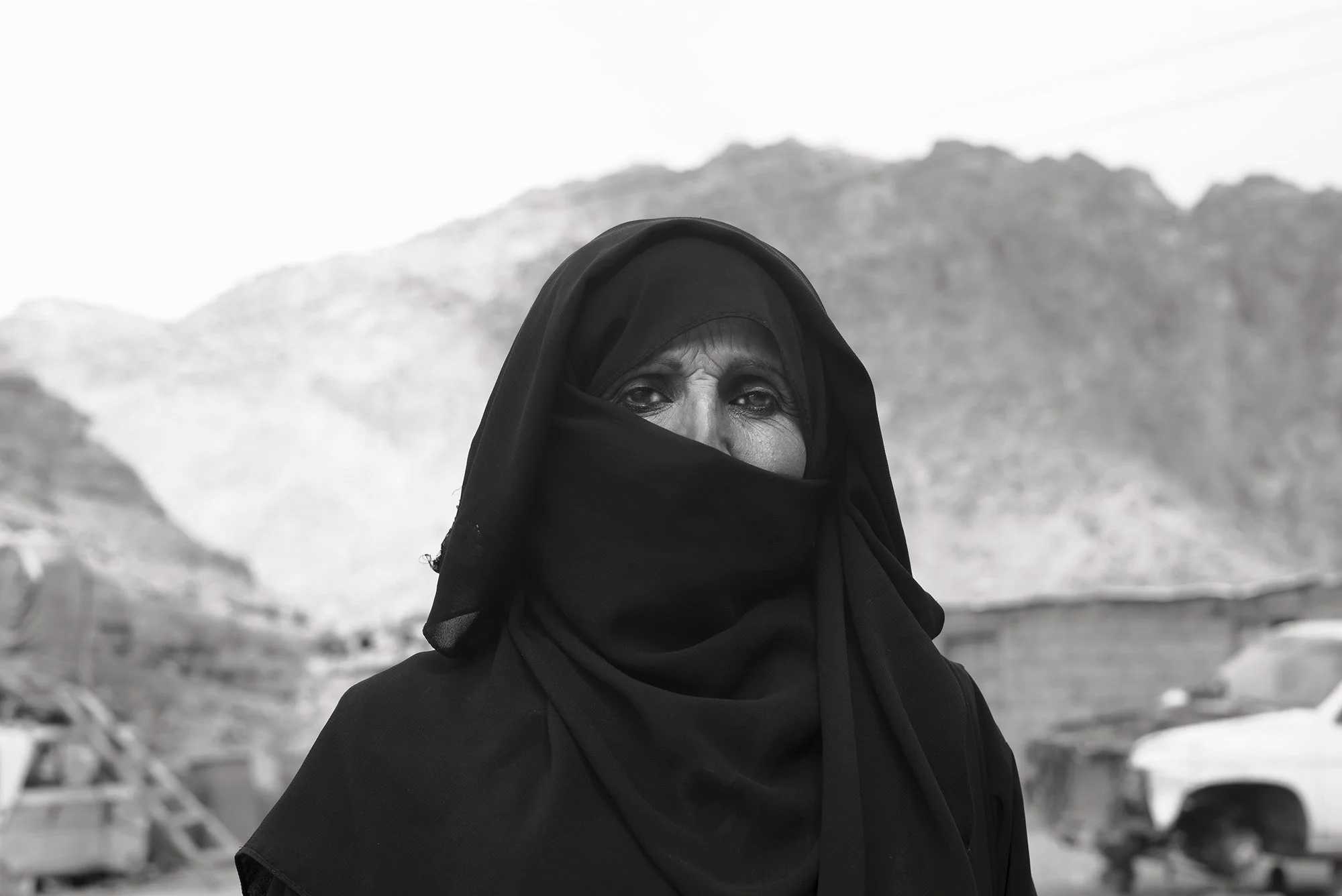 Close-up black and white portrait of a woman wearing a black niqab, with a blurred mountain landscape in the background.