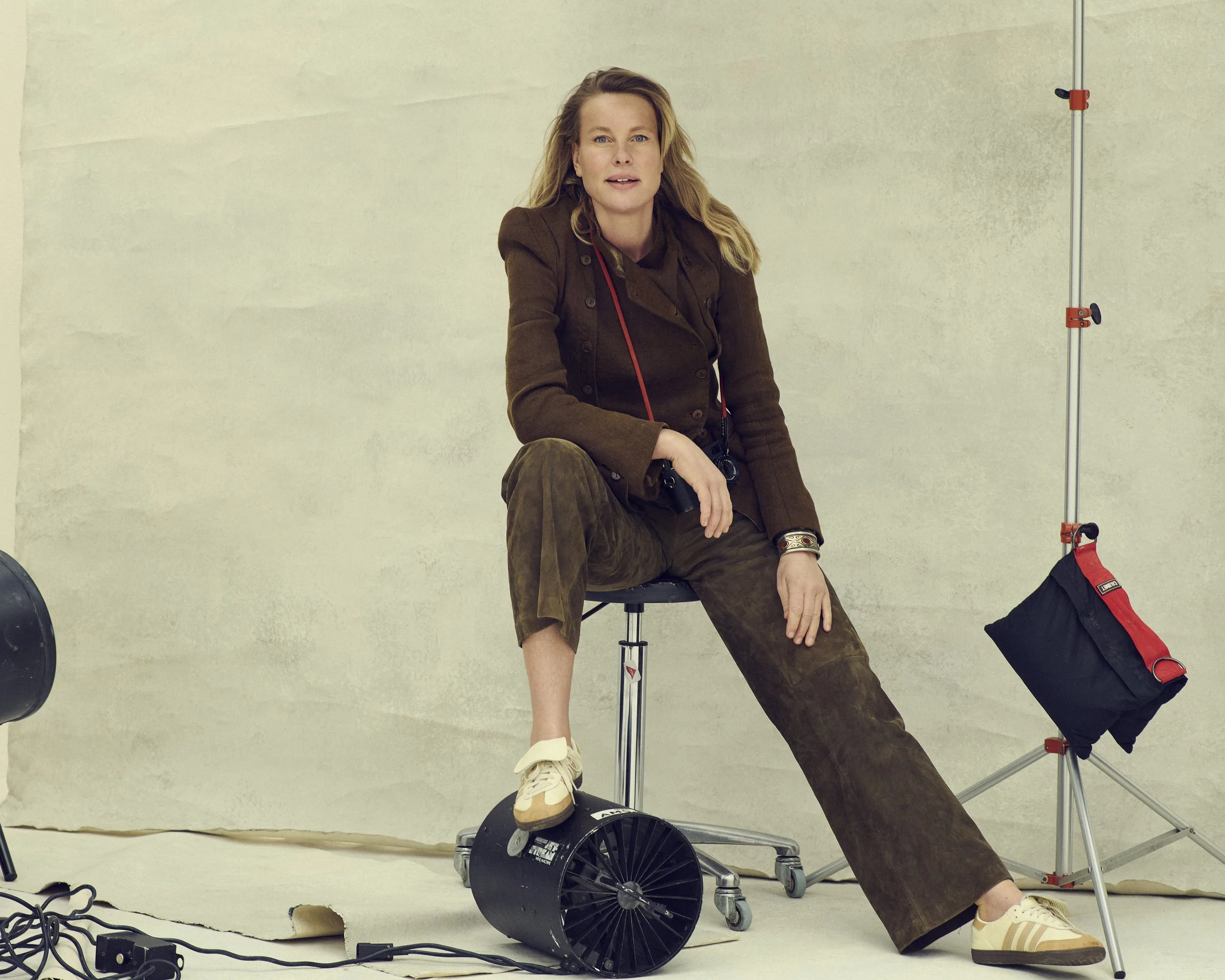 Professional portrait of photographer Isabelle Kettner sitting in her studio, wearing a brown jacket and trousers, with photography equipment in the background.