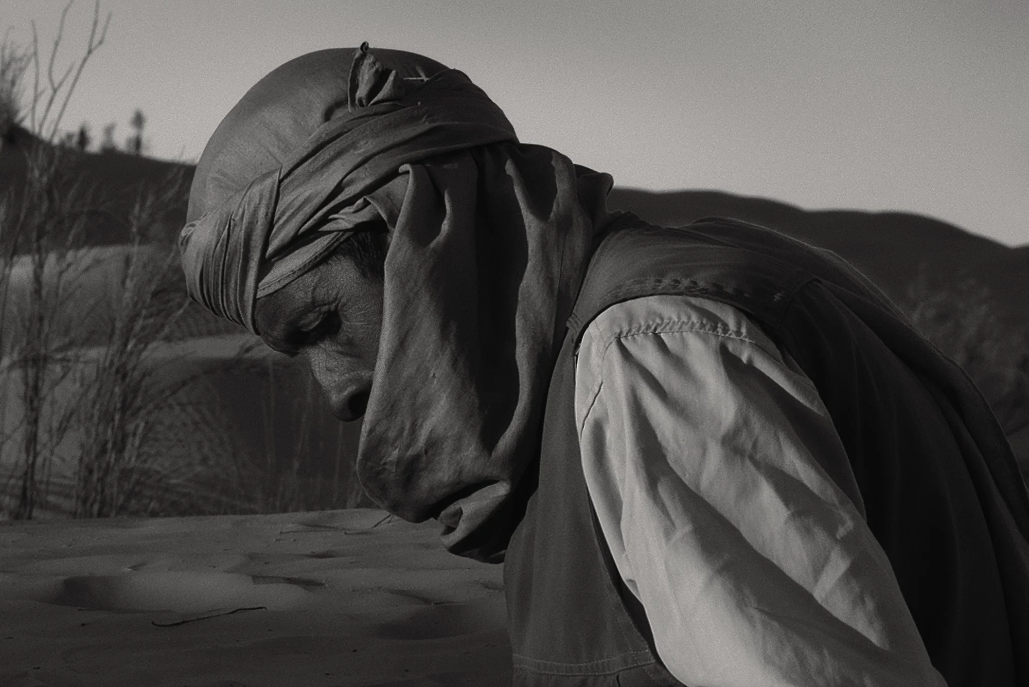 Intense black and white close-up profile of a man wearing a traditional headwrap (turban), looking down in a moment of reflection or prayer in the desert.