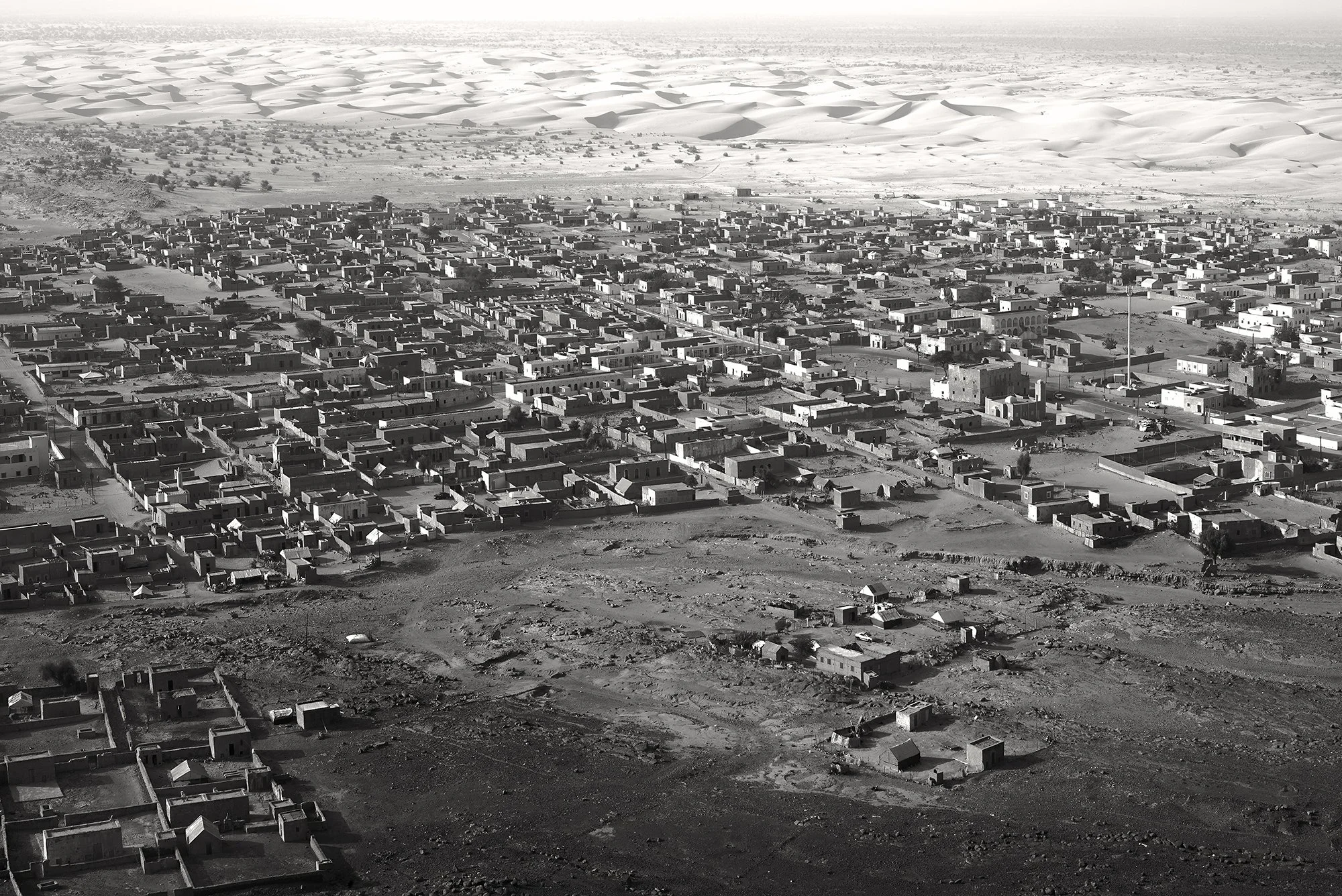 Aerial black and white view of the village Moudjiera, showing dense traditional architecture bordered by vast sand dunes.