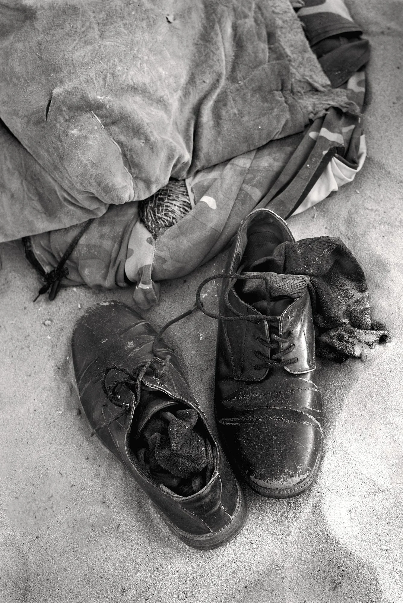 Still life monochrome photography of a pair of worn black dress shoes and socks resting on desert sand next to a heavy fabric bag.