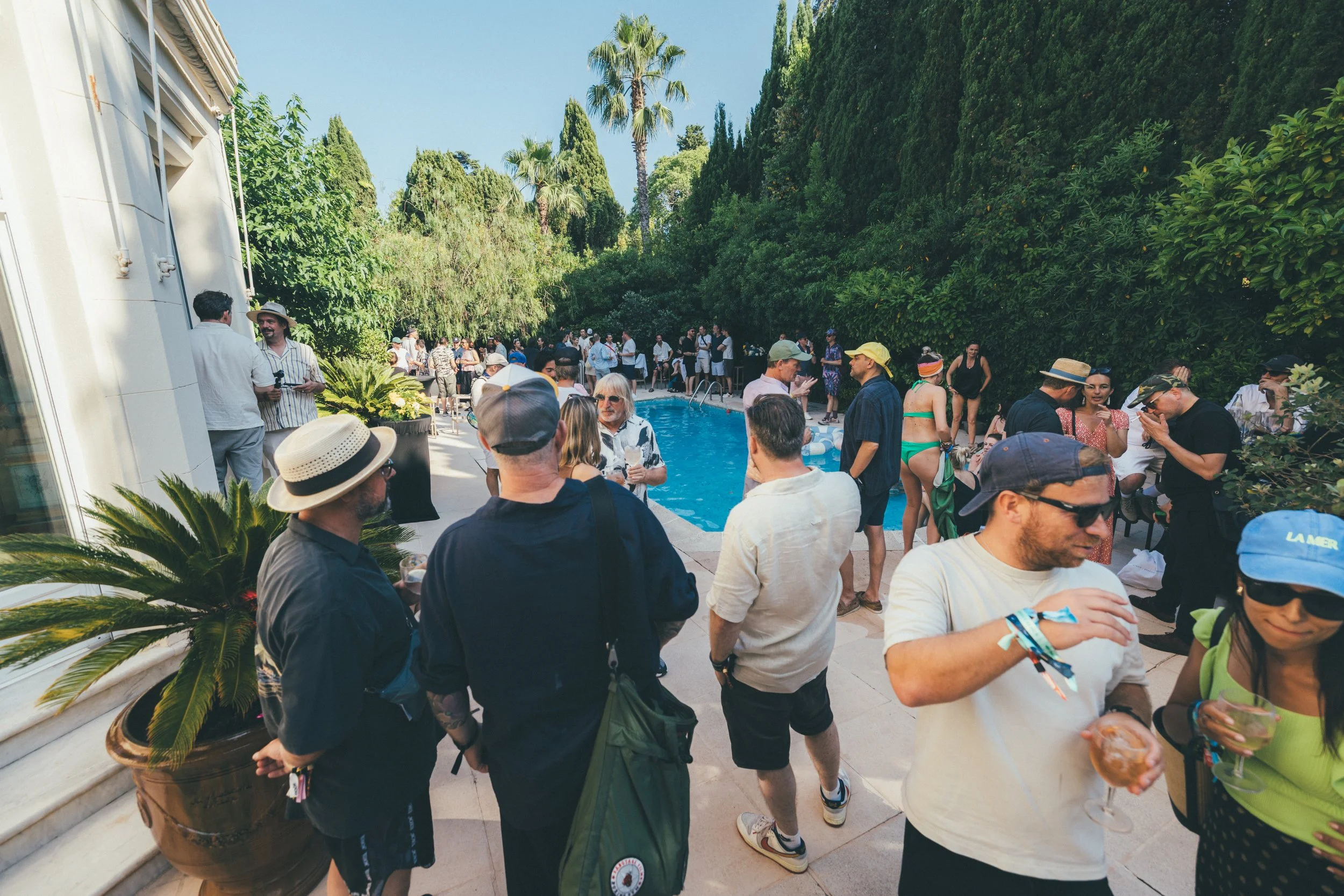 People gathering at a backyard pool party on a sunny day, with lush green trees in the background and some guests in swimsuits.