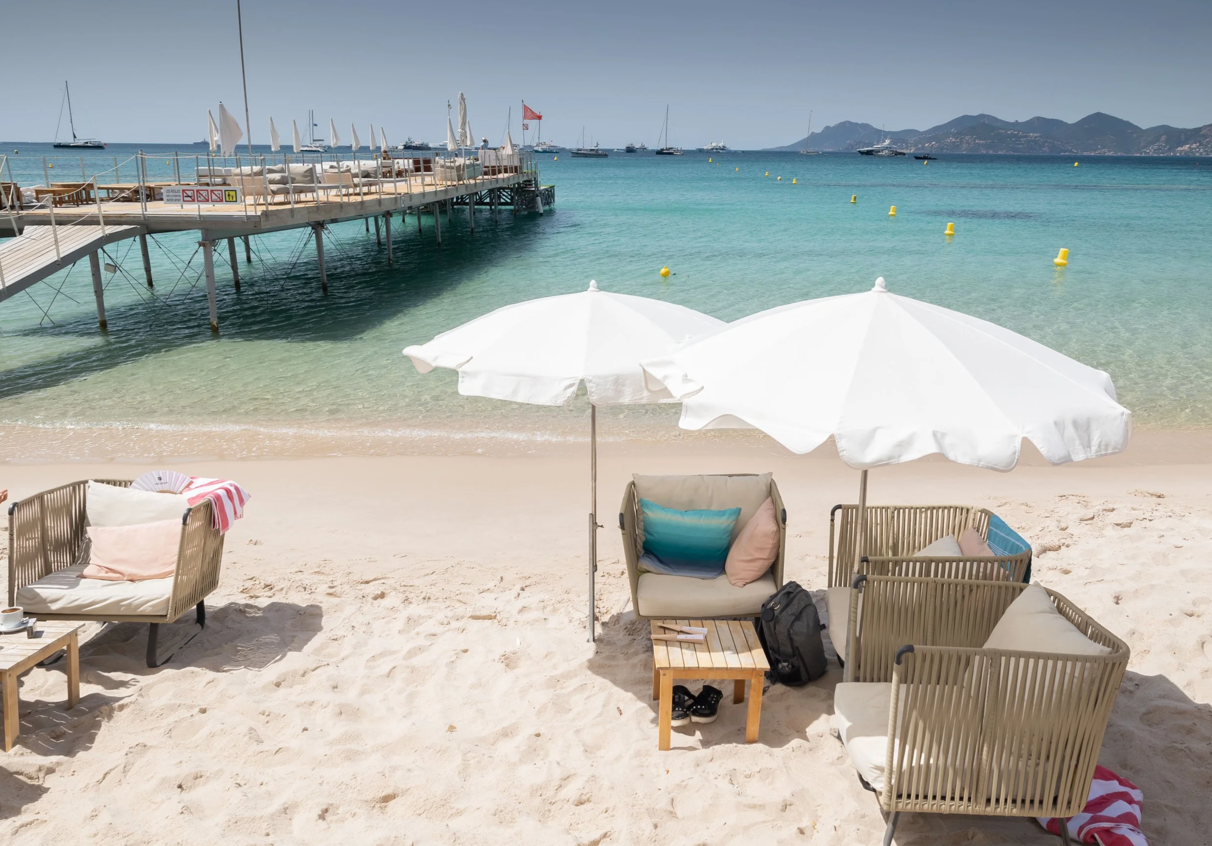 Beach with white umbrellas, outdoor seating, and a pier over turquoise water, with sailboats and mountains in the background.