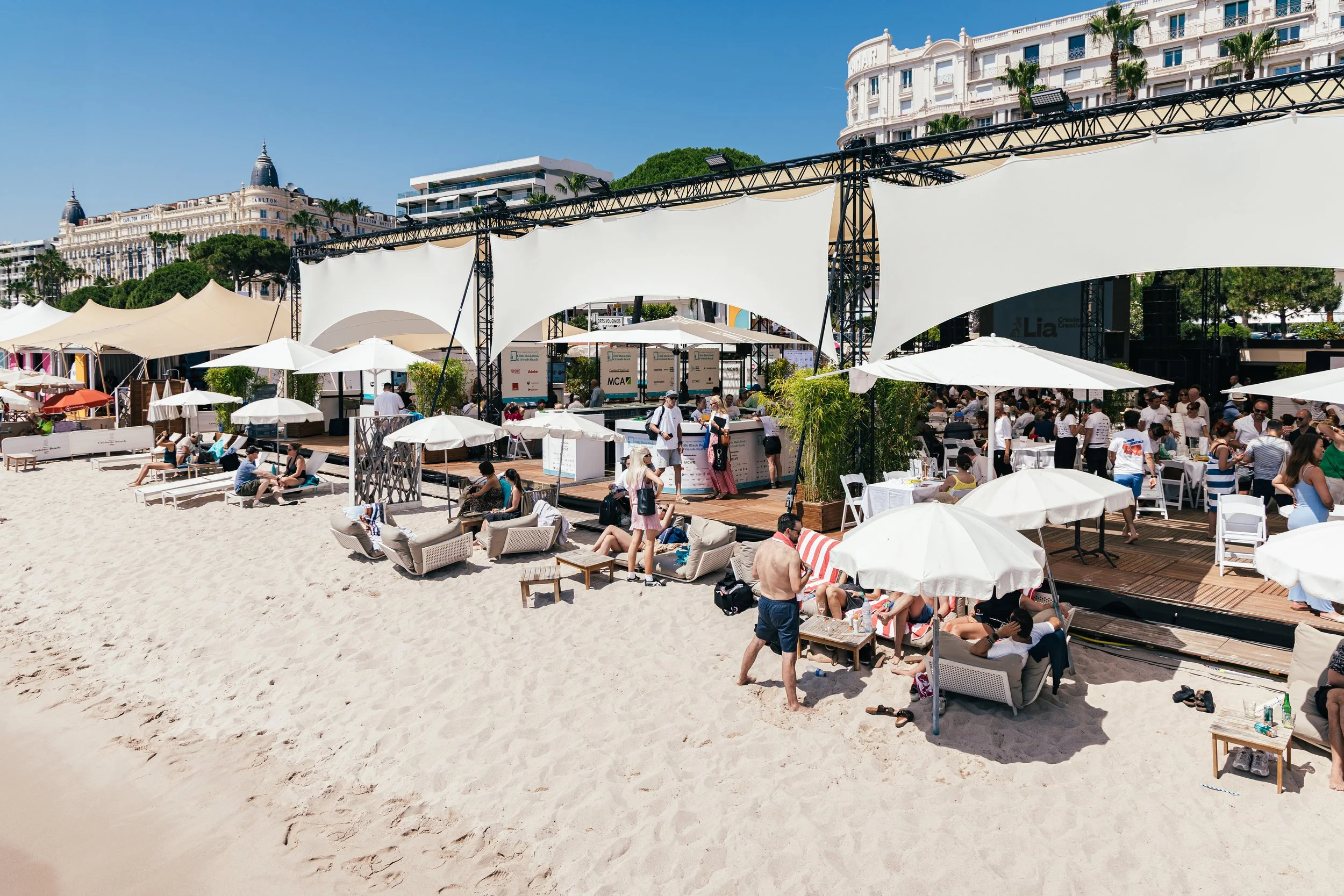 Beachside event with people relaxing under umbrellas and seated on lounge chairs, adjacent to a stage with white canopy coverings, in front of a row of hotel buildings and clear blue sky.