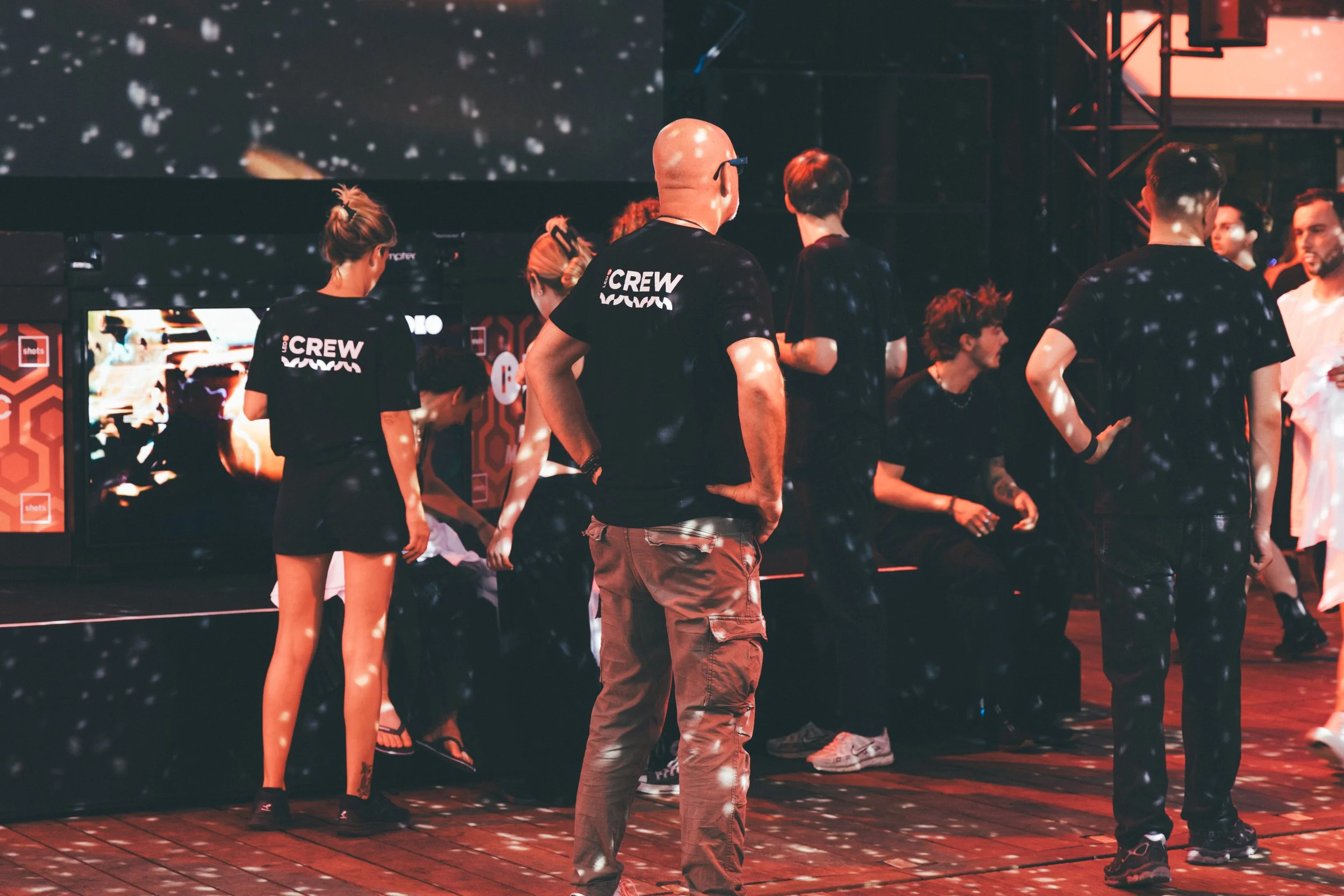 Group of people, some sitting and some standing, at a black and red event, with disco lighting effects on them and a large screen in the background.