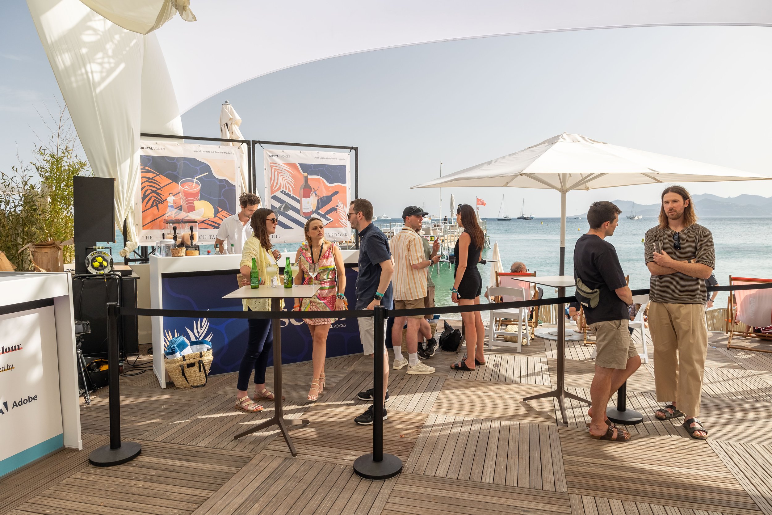 People socializing at a beachside bar under umbrellas with a body of water and boats in the background.