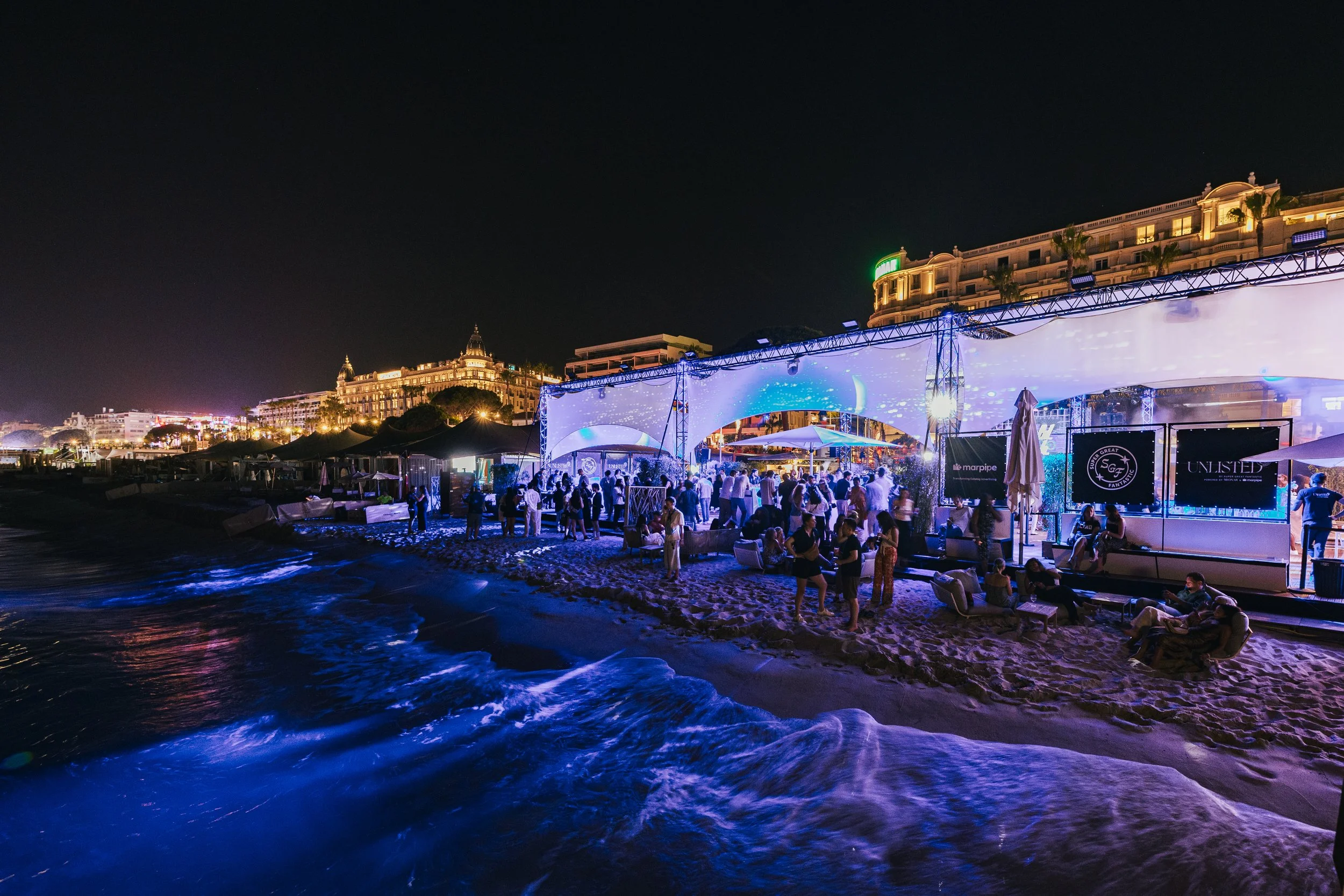 Nighttime beach scene with a lively party under a large illuminated tent, with people socializing and music. In the background, there are luxurious buildings and hotels along the shoreline.