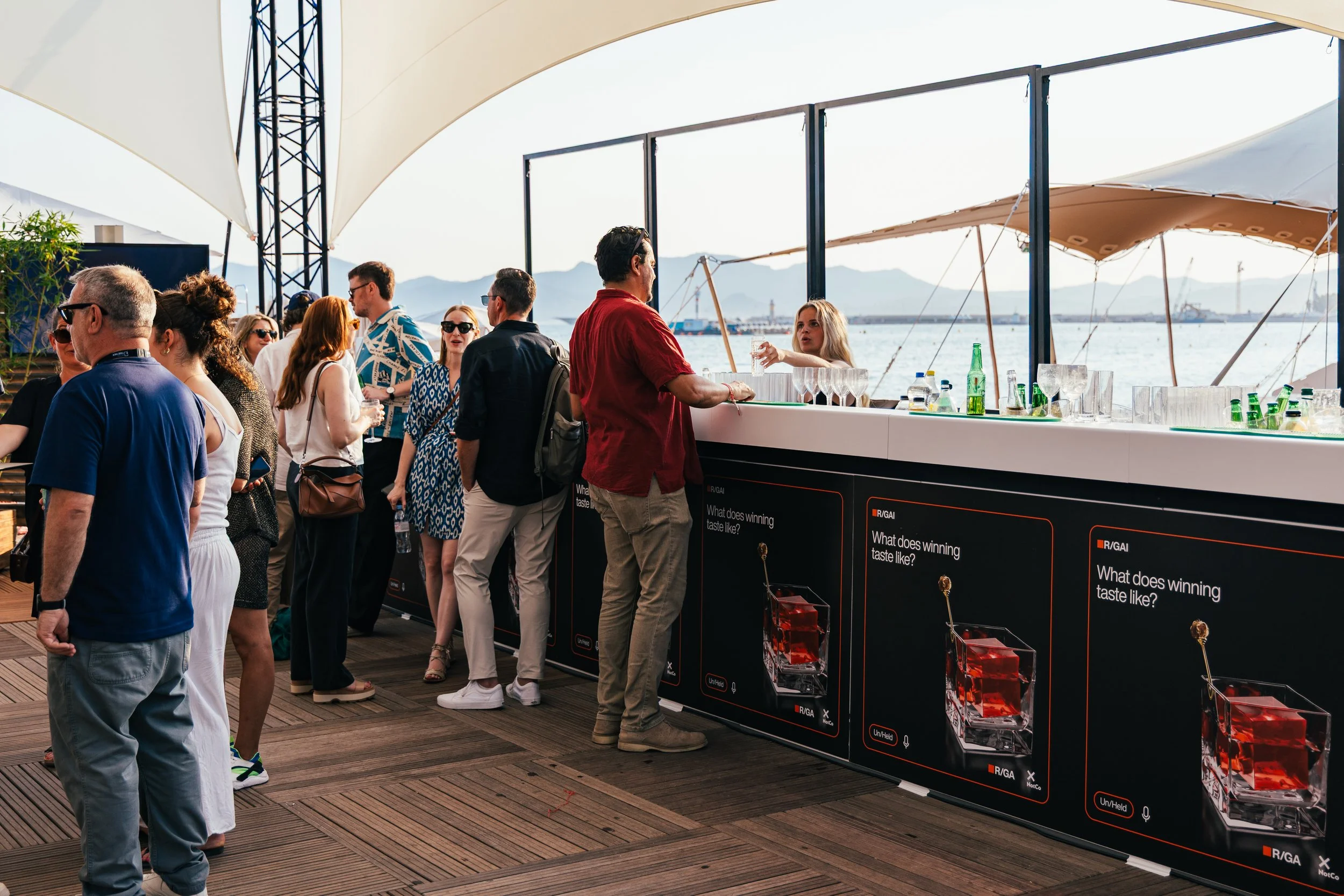 People gather at an outdoor bar near the water, with sailboats and mountains in the background, under a large tent.