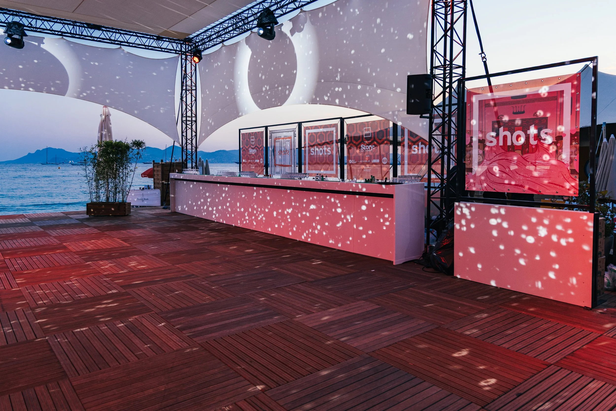 An outdoor event space with a wooden floor, a bar, and a large screen with the word 'shots' on it, set against a sunset over the sea with mountains in the background. There are stage lights and decorative light patterns on the surfaces.