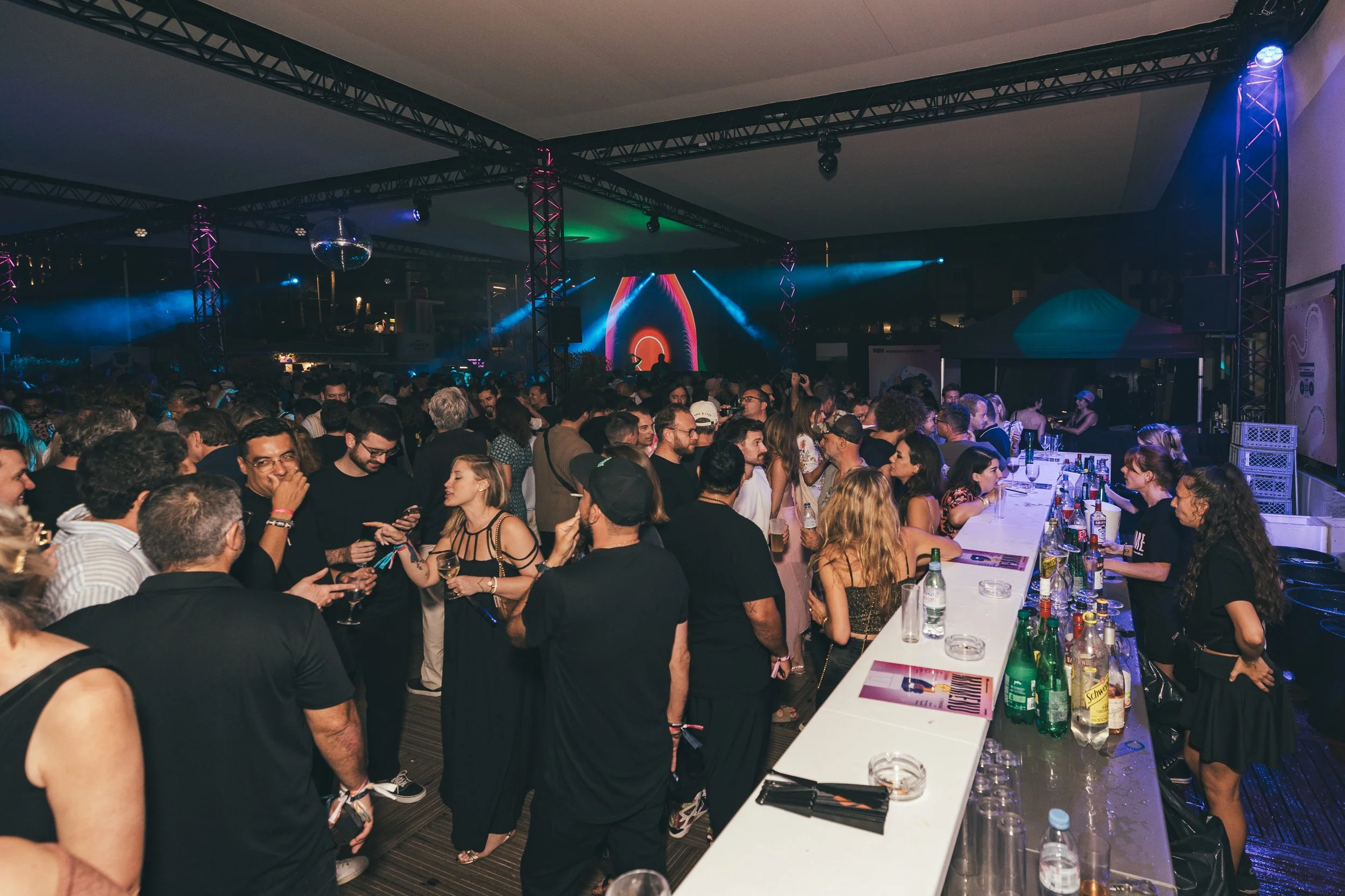 Crowd of people at a nightclub with a bar and LED screen, dancing and socializing under colorful lights.