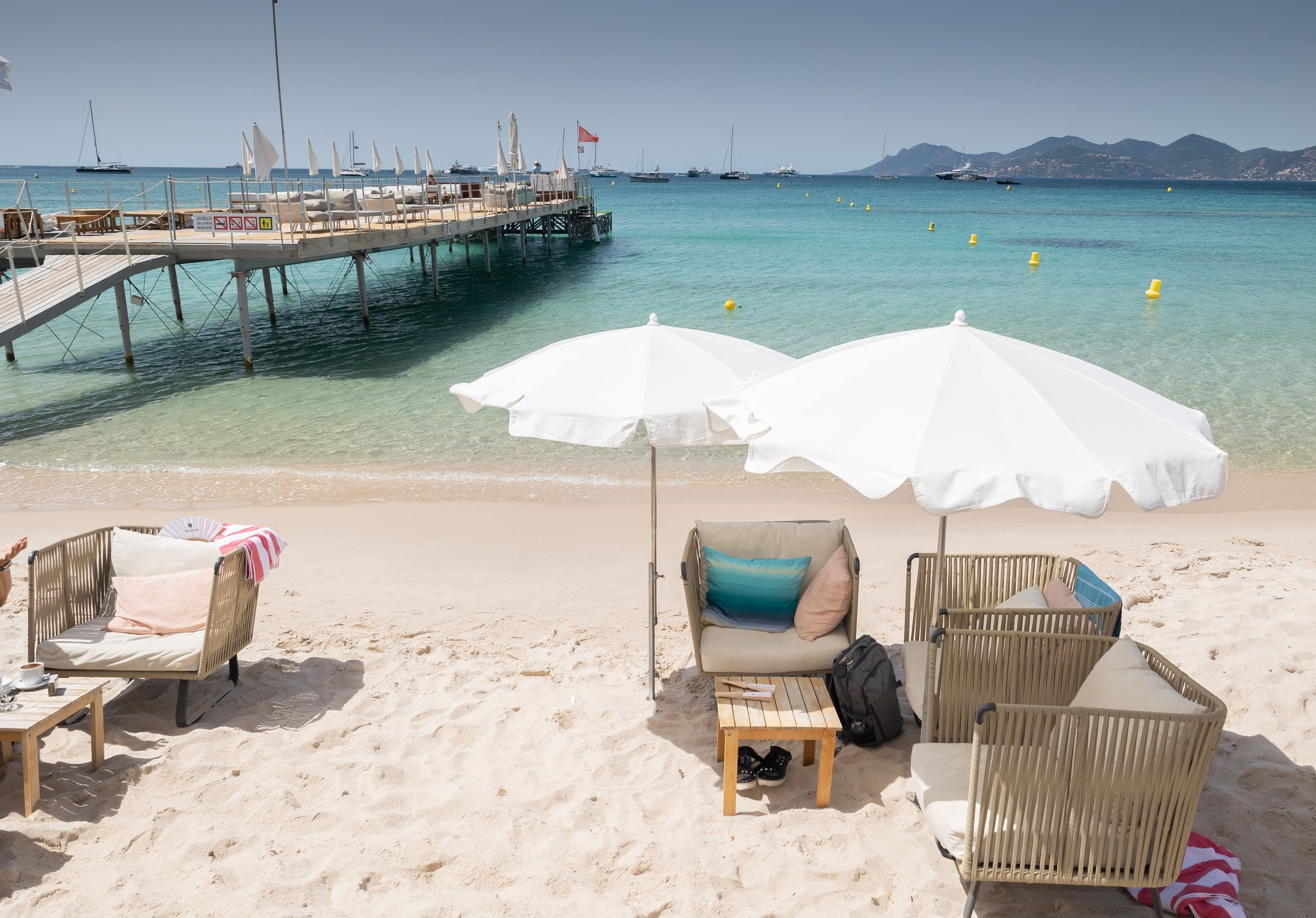 Beach with white umbrellas, outdoor seating, and a pier over turquoise water, with sailboats and mountains in the background.
