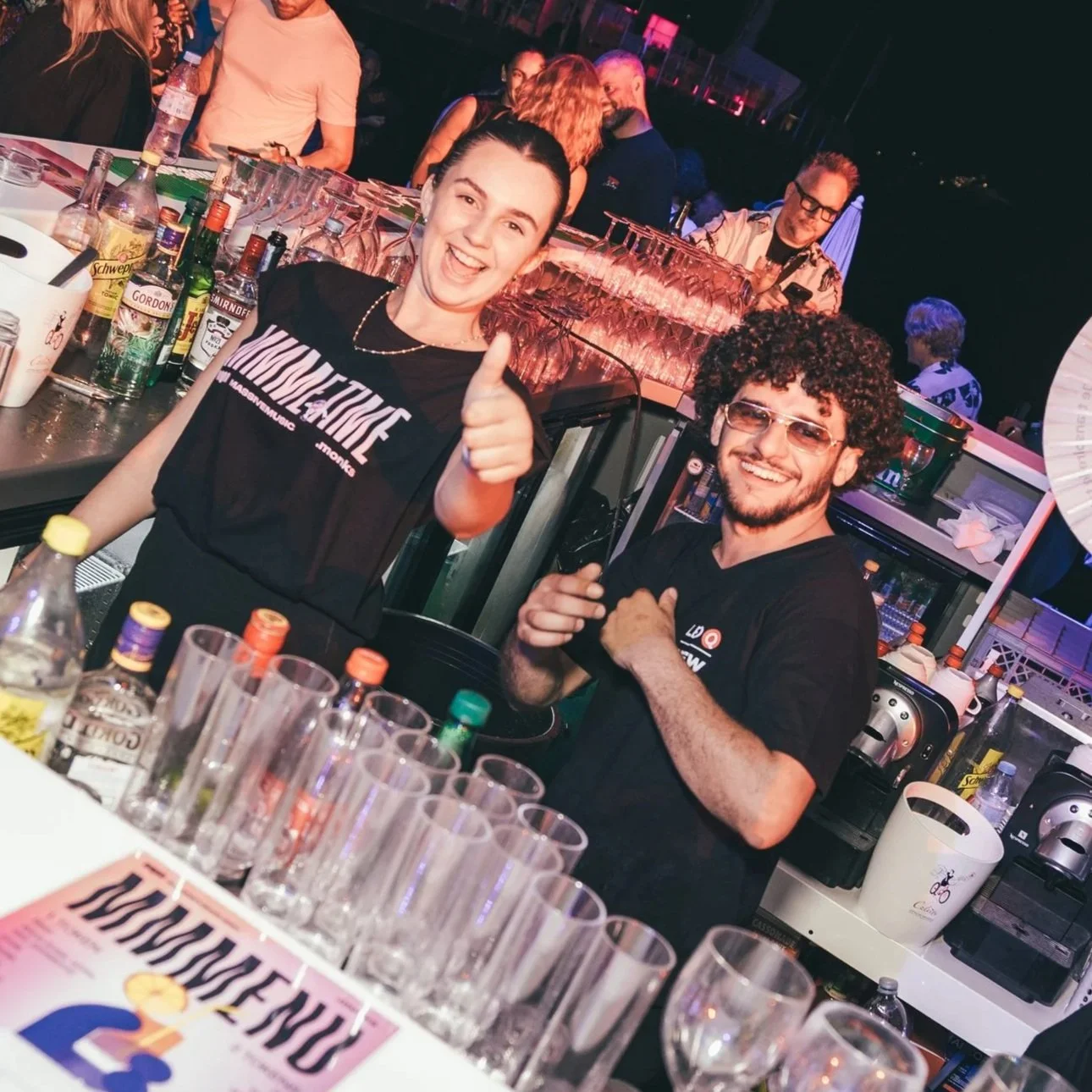 Two bartenders smiling at a bar, with one giving a thumbs-up, surrounded by glasses and bottles, in a lively nightclub or bar setting with people in the background.