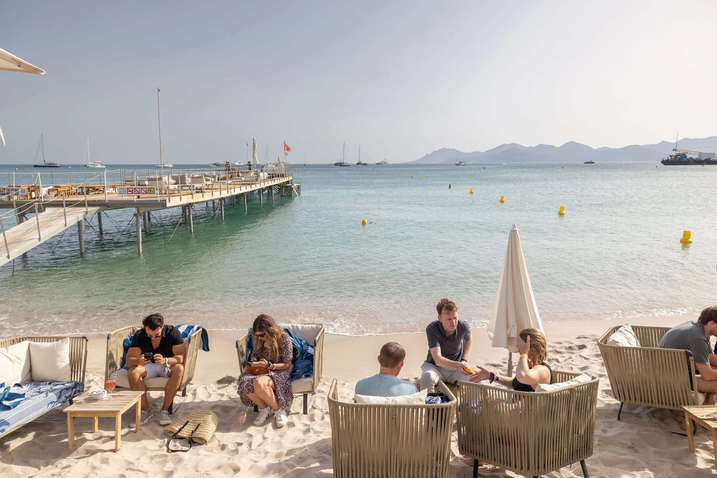 People relaxing on beach chairs and sofas on a sandy beach with a pier and boats in the water, mountains in the background.
